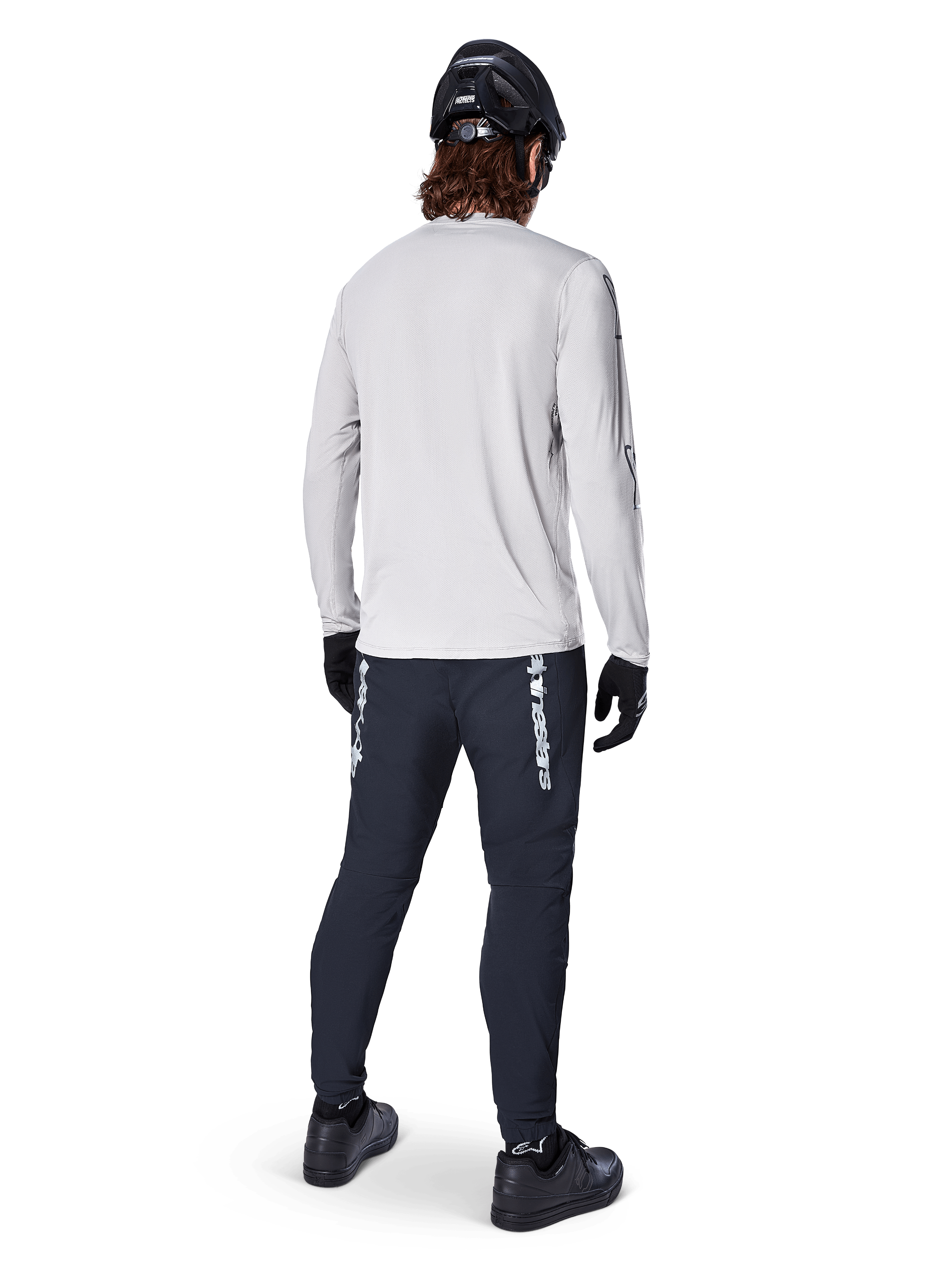 Alpinestars A-Dura Switch Jersey de manga larga, gris claro, vista posterior de un ciclista con pantalones de ciclismo negros, casco y guantes, equipamiento de alto rendimiento para MTB con parte trasera más larga