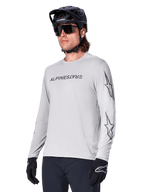 Alpinestars A-Dura Switch Long Sleeve Jersey, camiseta de manga larga para bicicleta de montaña, gris claro, serigrafía negra de la marca en el pecho y logotipos de estrellas en las mangas, usado con casco negro de MTB, gafas de sol oscuras y guantes negros, equipamiento deportivo para ciclismo off-road