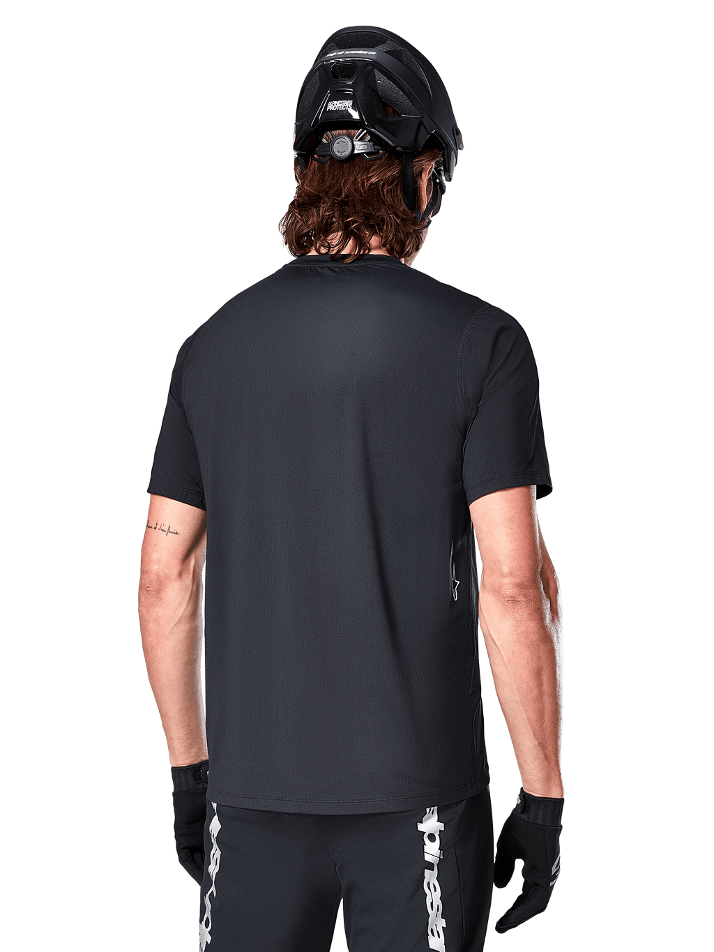 Jersey de manga corta Alpinestars A-Dura Switch, negro, vista trasera en un modelo con casco de ciclismo negro y pantalones cortos de MTB, destacando el ajuste regular y la cola extendida diseñada para el ciclismo de montaña.