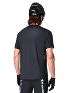 Jersey de manga corta Alpinestars A-Dura Switch, negro, vista trasera en un modelo con casco de ciclismo negro y pantalones cortos de MTB, destacando el ajuste regular y la cola extendida diseñada para el ciclismo de montaña.