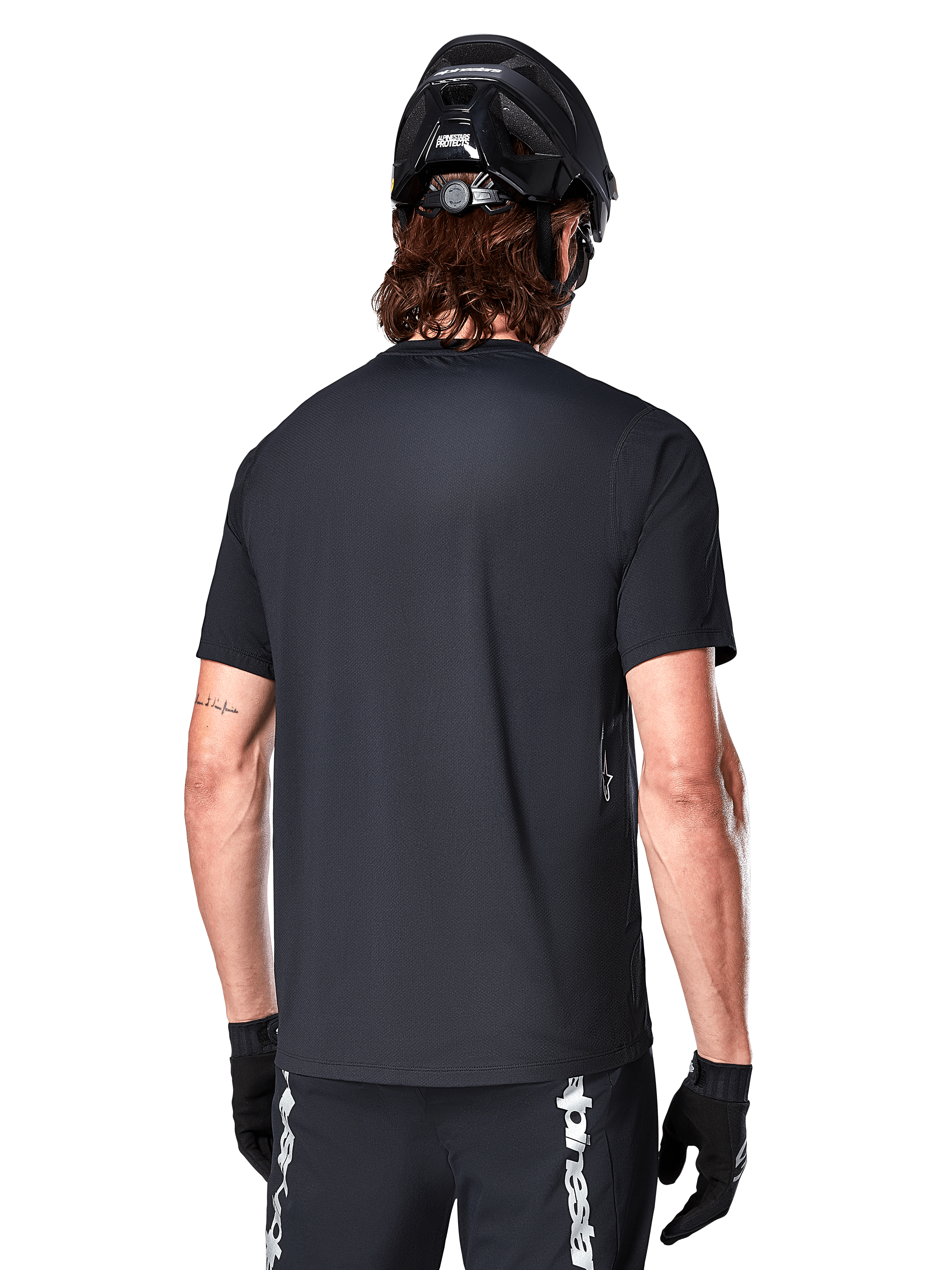 Jersey de manga corta Alpinestars A-Dura Switch, negro, vista trasera en un modelo con casco de ciclismo negro y pantalones cortos de MTB, destacando el ajuste regular y la cola extendida diseñada para el ciclismo de montaña.