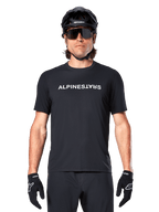 Ciclista de montaña con el jersey de manga corta Alpinestars A-Dura Switch en color negro, con un logotipo de la marca en blanco en el pecho, combinado con casco negro, gafas de sol grandes y guantes negros, ropa de ciclismo profesional para trail.