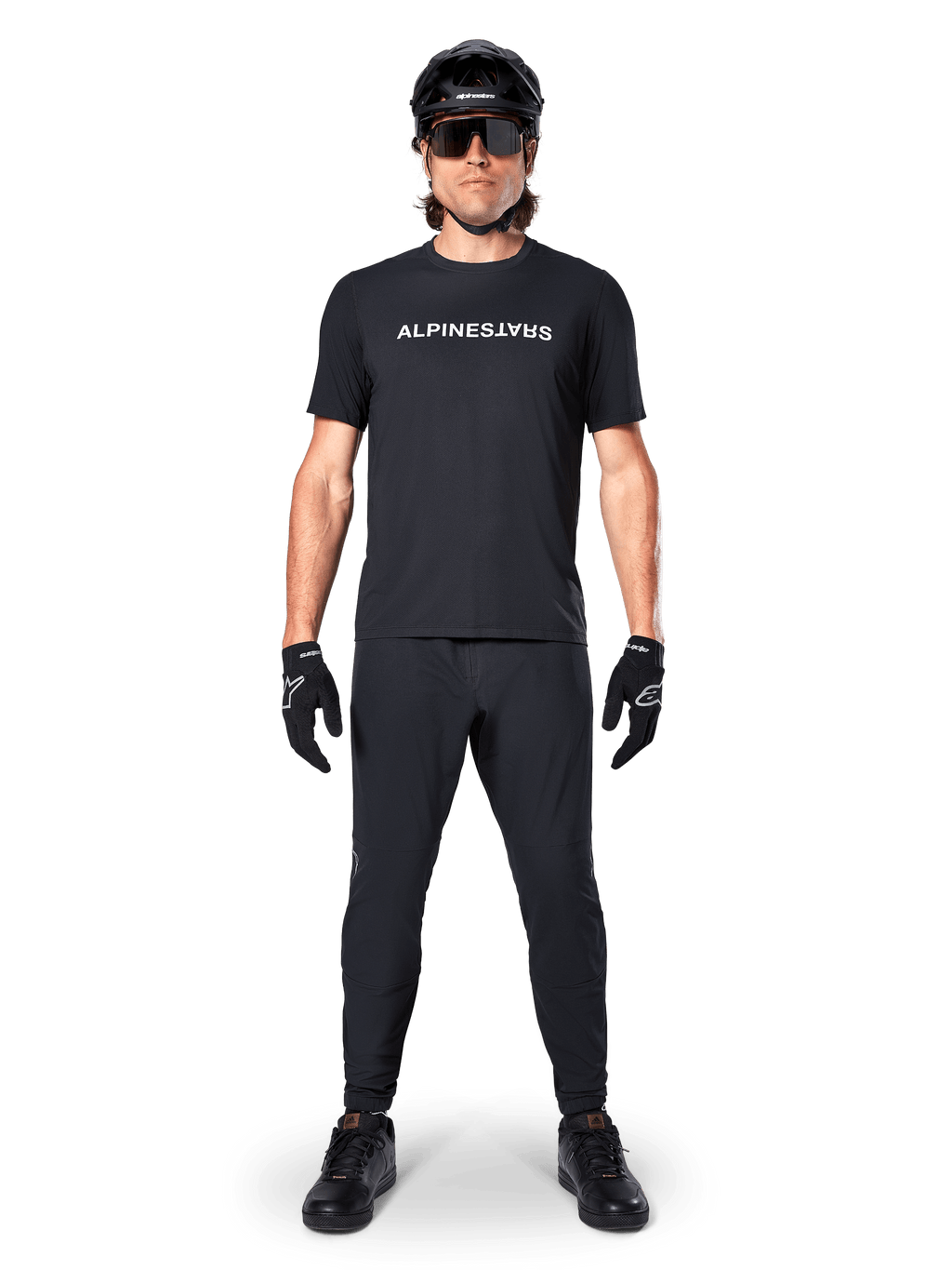 Ciclista de montaña con el jersey de manga corta Alpinestars A-Dura Switch en negro, con un logotipo blanco estilizado en el pecho, combinado con pantalones de MTB negros, casco y guantes, equipamiento de ciclismo completo listo para el sendero.