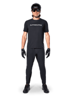 Ciclista de montaña con el jersey de manga corta Alpinestars A-Dura Switch en negro, con un logotipo blanco estilizado en el pecho, combinado con pantalones de MTB negros, casco y guantes, equipamiento de ciclismo completo listo para el sendero.