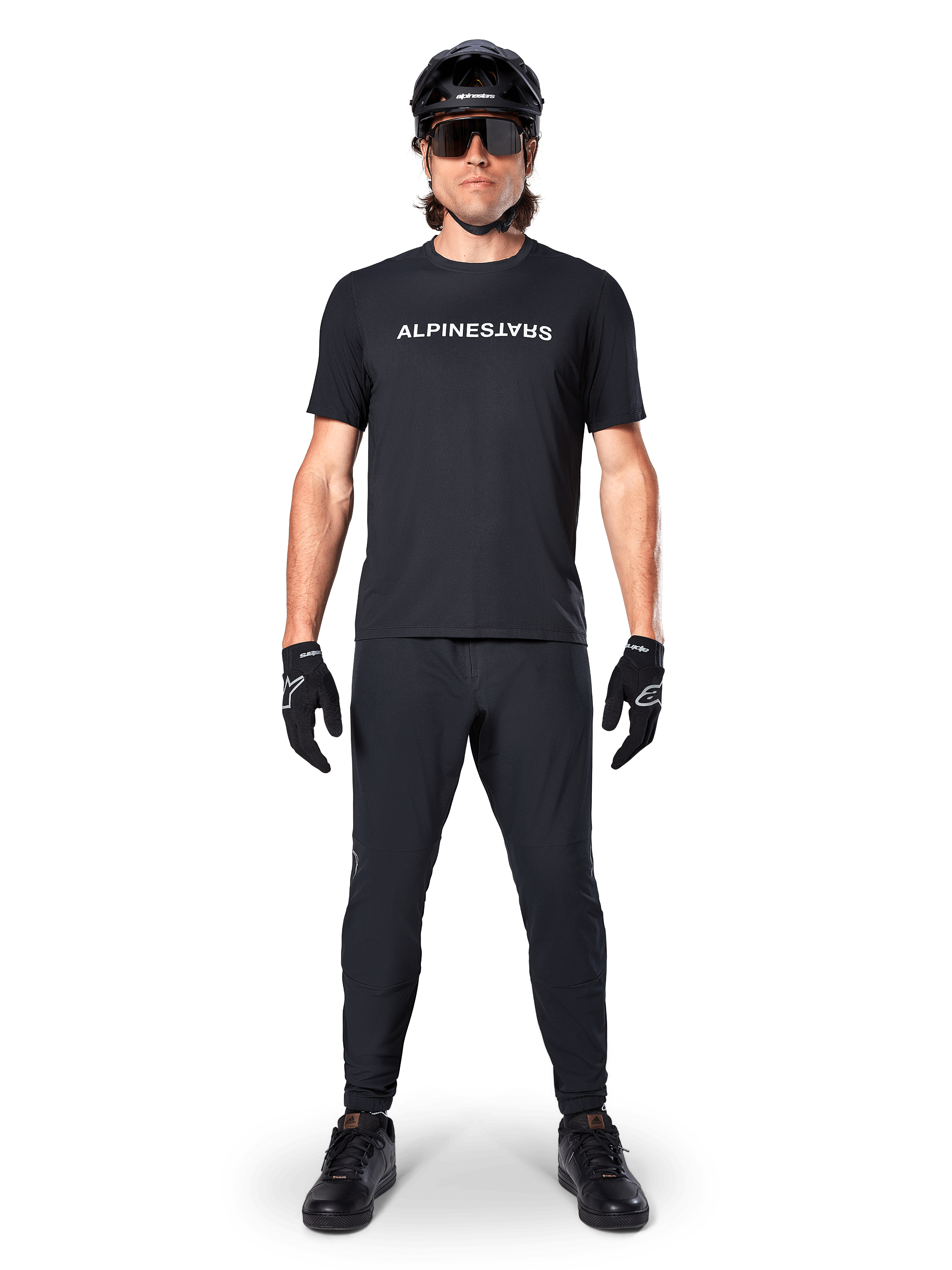 Ciclista de montaña con el jersey de manga corta Alpinestars A-Dura Switch en negro, con un logotipo blanco estilizado en el pecho, combinado con pantalones de MTB negros, casco y guantes, equipamiento de ciclismo completo listo para el sendero.