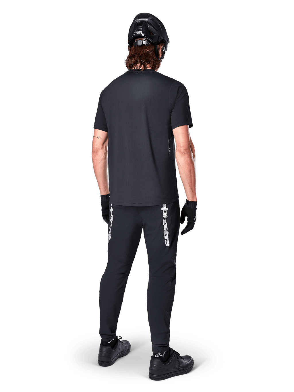 Jersey Alpinestars A-Dura Switch - Manga Corta, negro, vista trasera de un ciclista con equipamiento completo de bicicleta de montaña, incluyendo pantalones negros a juego con marca denominativa blanca, casco negro y guantes, ropa de ciclismo de alto rendimiento para trail