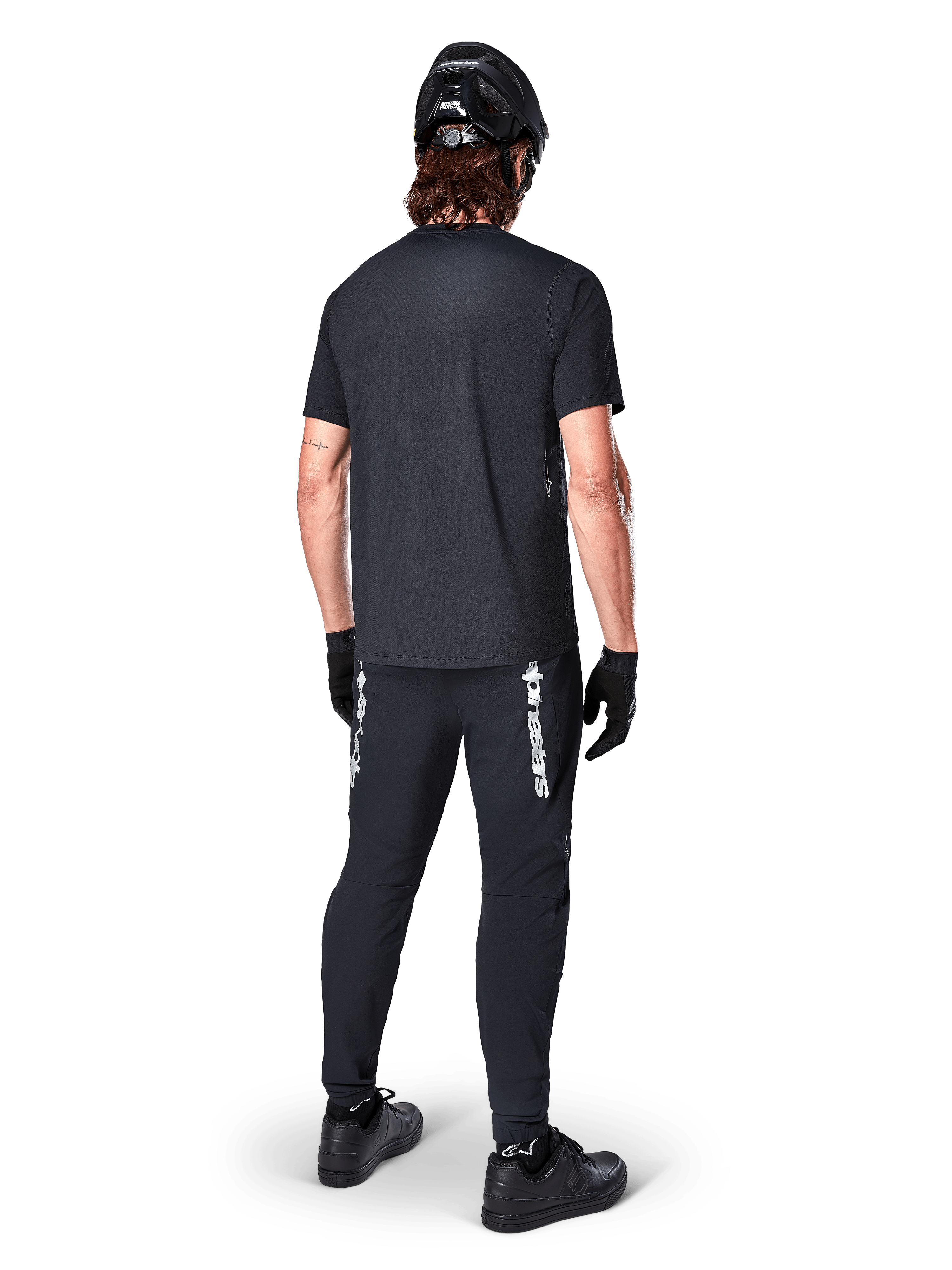 Jersey Alpinestars A-Dura Switch - Manga Corta, negro, vista trasera de un ciclista con equipamiento completo de bicicleta de montaña, incluyendo pantalones negros a juego con marca denominativa blanca, casco negro y guantes, ropa de ciclismo de alto rendimiento para trail