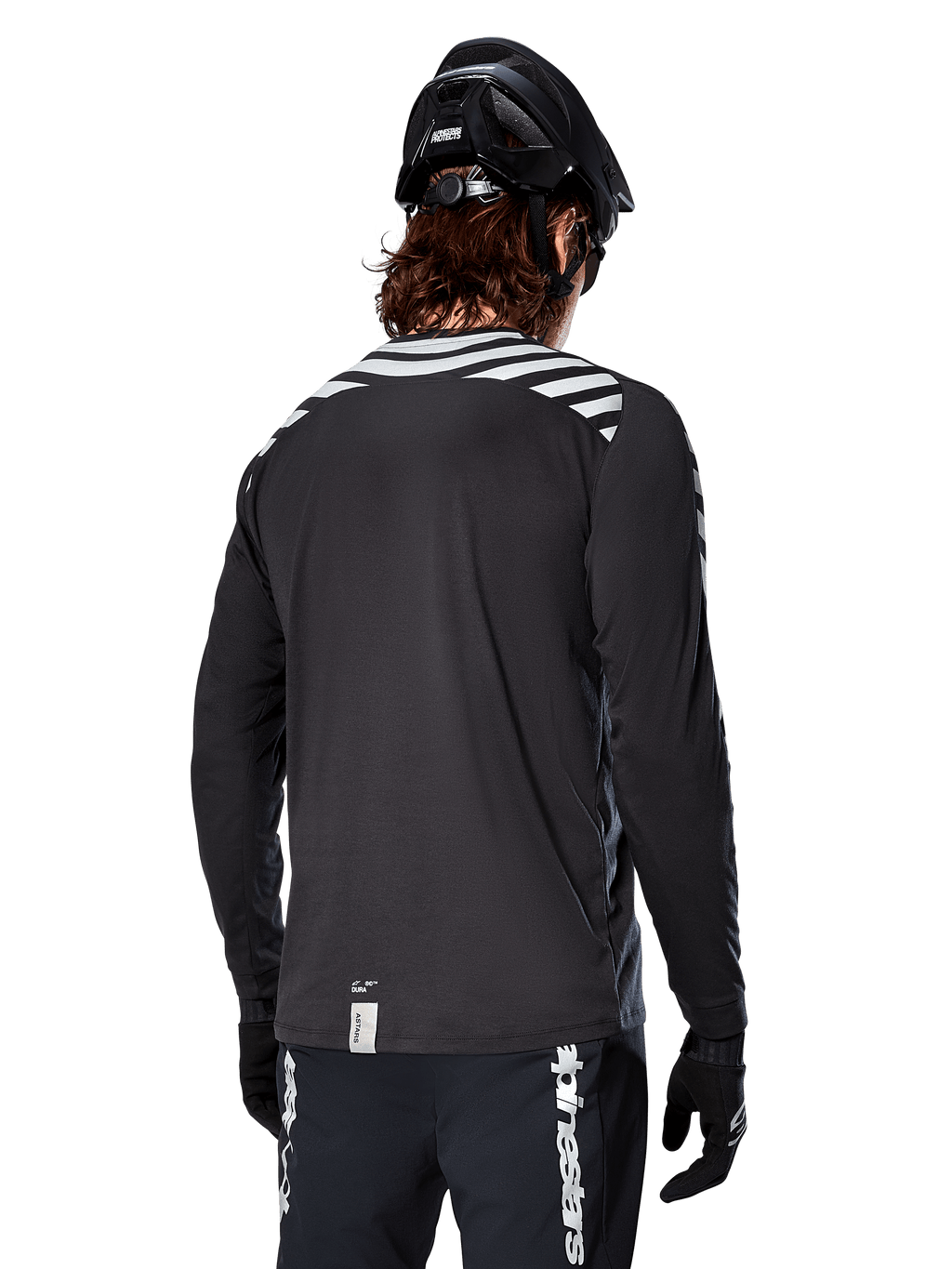 Jersey de manga larga Alpinestars A-Dura Dri Oscar, negro reflectante, vista trasera que muestra franjas diagonales reflectantes en los hombros y una parte trasera más larga, usado con casco y guantes de MTB negros, equipamiento técnico para ciclismo de montaña