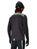 Jersey de manga larga Alpinestars A-Dura Dri Oscar, negro reflectante, vista trasera que muestra franjas diagonales reflectantes en los hombros y una parte trasera más larga, usado con casco y guantes de MTB negros, equipamiento técnico para ciclismo de montaña