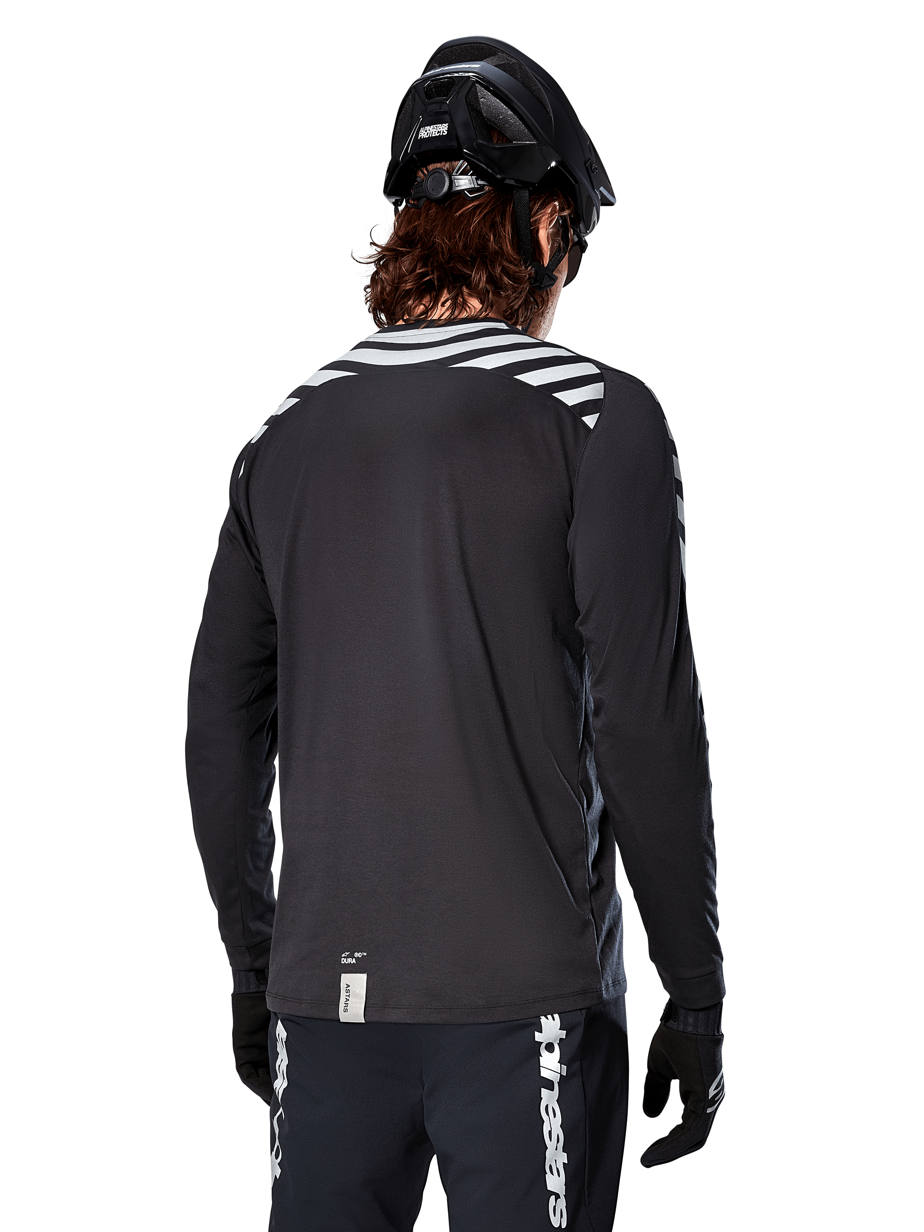 Jersey de manga larga Alpinestars A-Dura Dri Oscar, negro reflectante, vista trasera que muestra franjas diagonales reflectantes en los hombros y una parte trasera más larga, usado con casco y guantes de MTB negros, equipamiento técnico para ciclismo de montaña