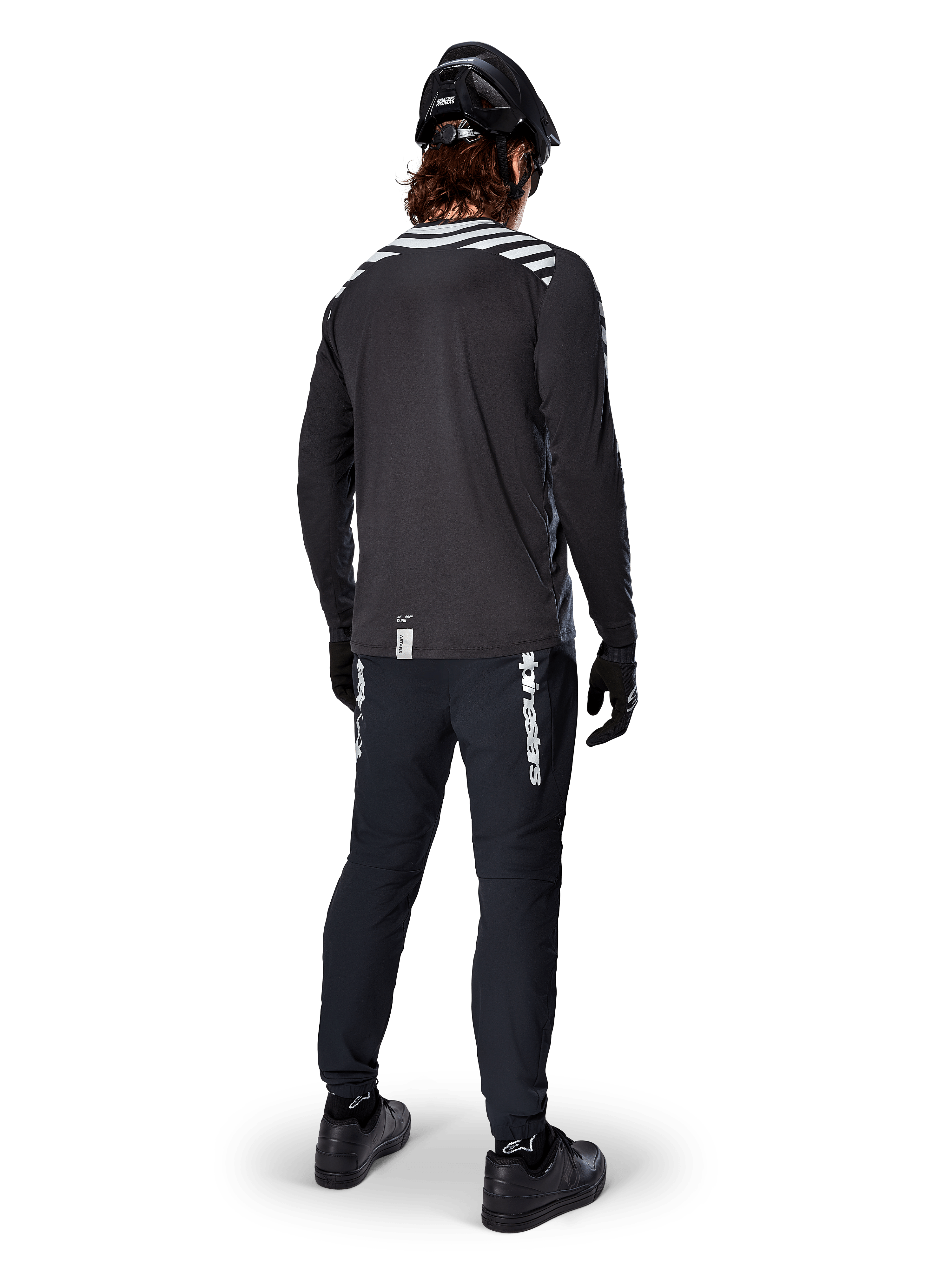 Ciclista de montaña vistiendo el Jersey de Manga Larga Alpinestars A-Dura Dri Oscar en Negro Reflectante, visto desde atrás, combinado con pantalones técnicos negros, casco y guantes, con gráficos de rayas reflectantes en los hombros y una parte trasera más larga para ciclismo de montaña.