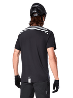 Jersey Alpinestars A-Dura Dri Oscar, jersey de MTB de manga corta, negro reflectante, vista trasera de un modelo con casco de ciclismo y guantes, con rayas grises reflectantes en los hombros y parte trasera caída, tejido técnico transpirable para ciclismo de montaña