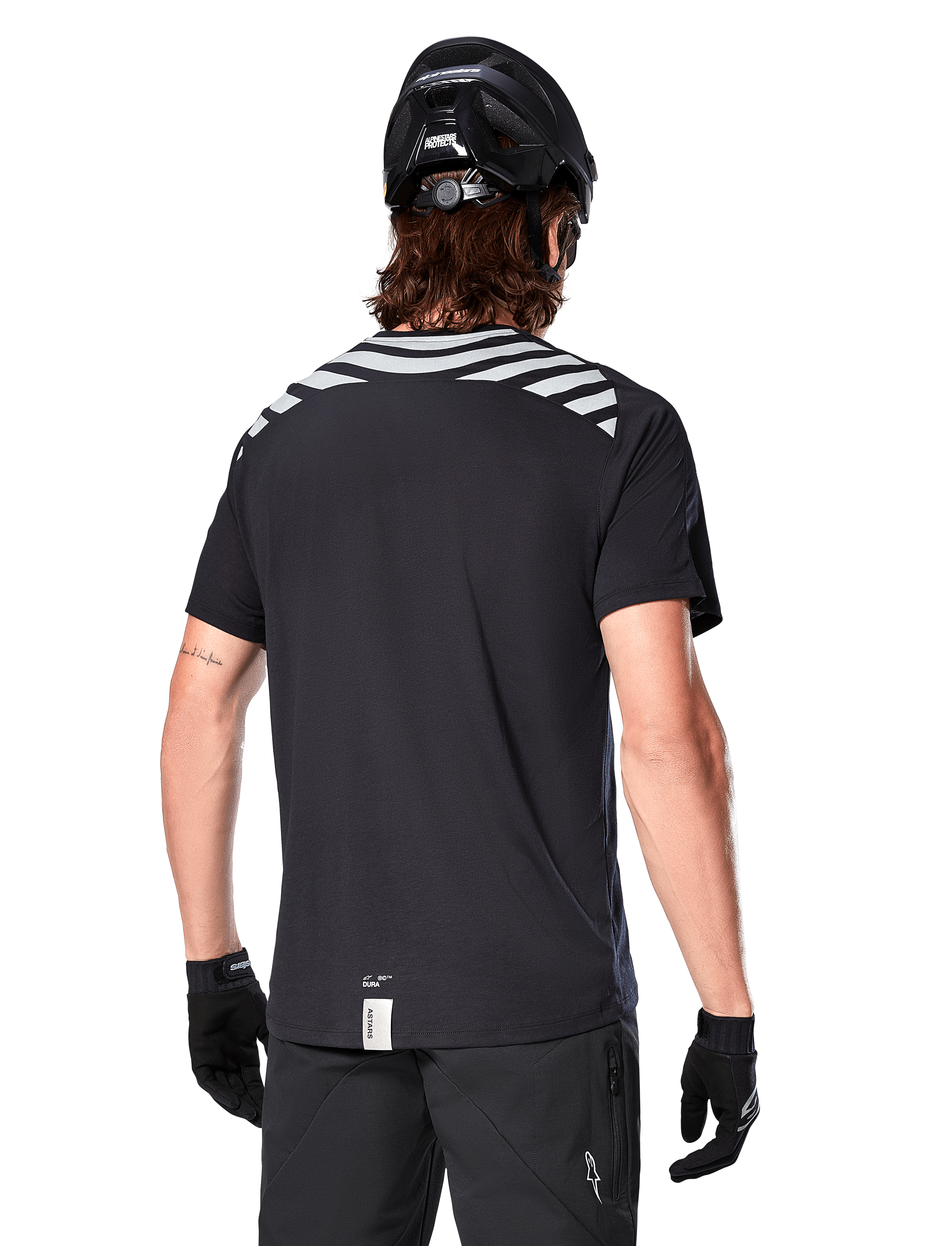 Jersey Alpinestars A-Dura Dri Oscar, jersey de MTB de manga corta, negro reflectante, vista trasera de un modelo con casco de ciclismo y guantes, con rayas grises reflectantes en los hombros y parte trasera caída, tejido técnico transpirable para ciclismo de montaña