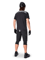 Jersey de manga corta Alpinestars A-Dura Dri Oscar, negro reflectante, vista trasera de un ciclista usando el jersey con pantalones cortos de montaña negros, casco y rodilleras, con gráficos reflectantes en los hombros y diseño de cola caída para MTB.
