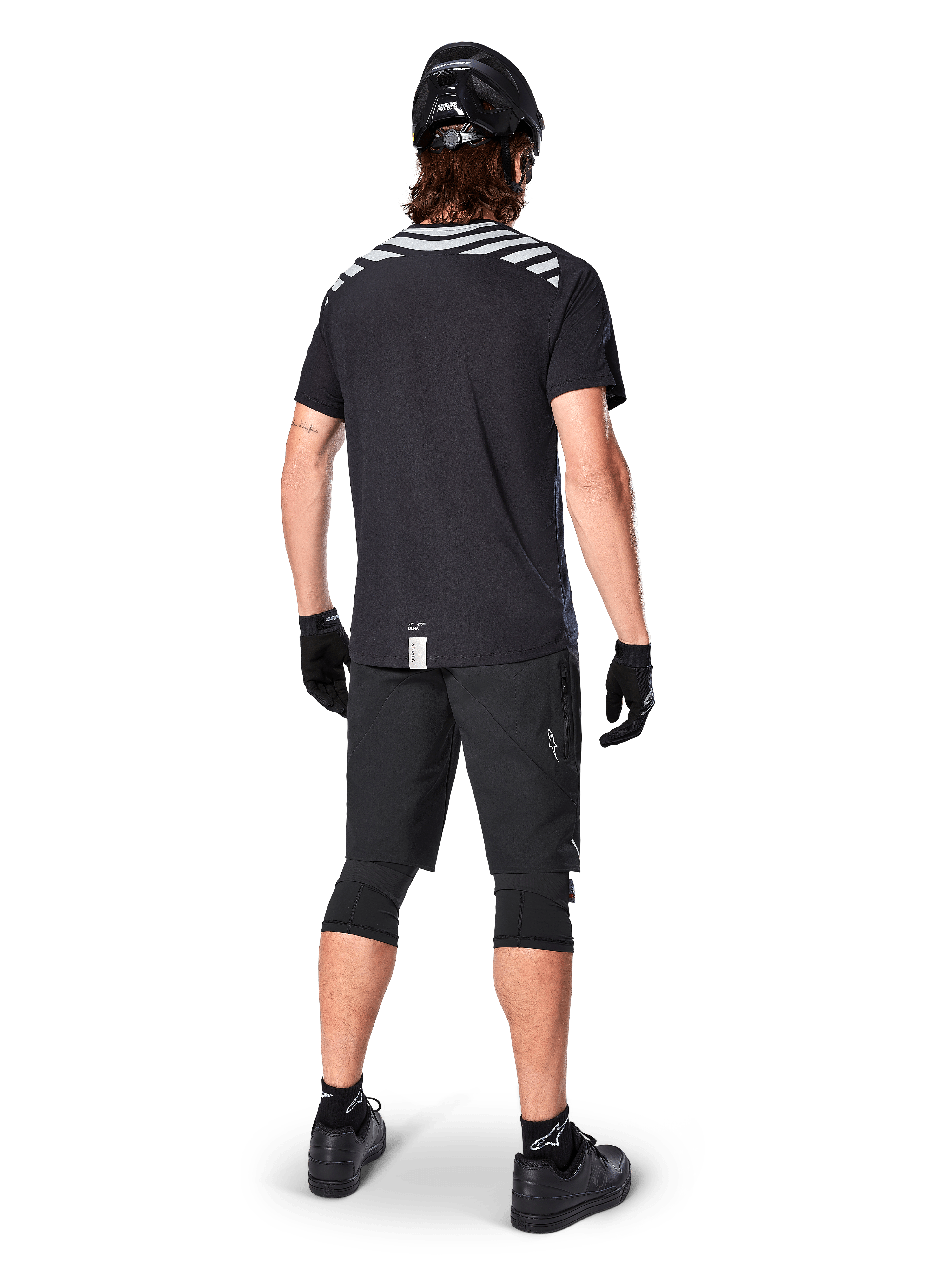 Jersey de manga corta Alpinestars A-Dura Dri Oscar, negro reflectante, vista trasera de un ciclista usando el jersey con pantalones cortos de montaña negros, casco y rodilleras, con gráficos reflectantes en los hombros y diseño de cola caída para MTB.