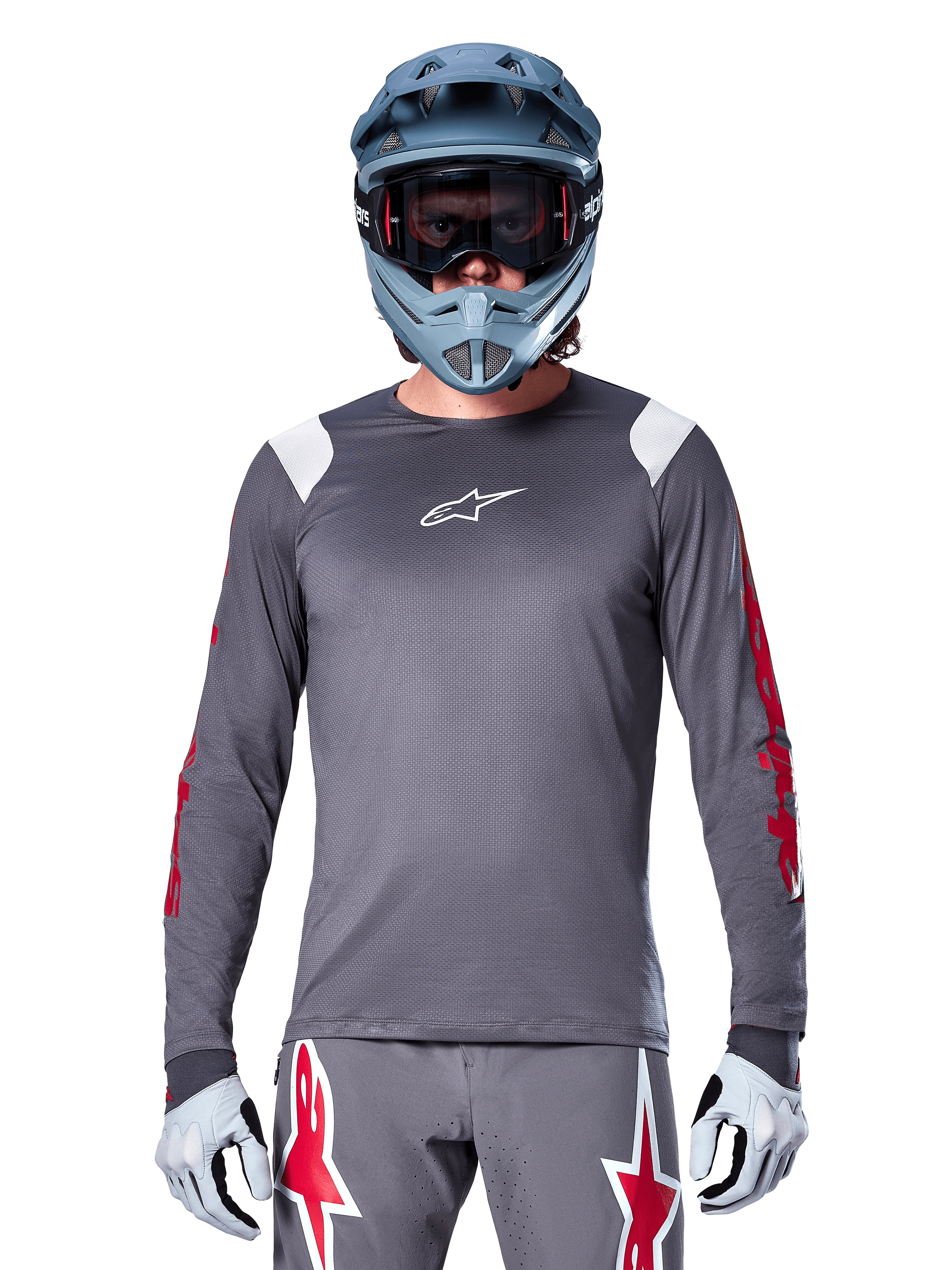 Ciclista de MTB con jersey de manga larga Alpinestars A-Supra Astar, gris oscuro con gráficos rojos en las mangas y detalles blancos en los hombros, pantalones grises a juego con logotipos de estrellas rojas, casco integral azul y gafas, equipamiento coordinado para ciclismo todoterreno