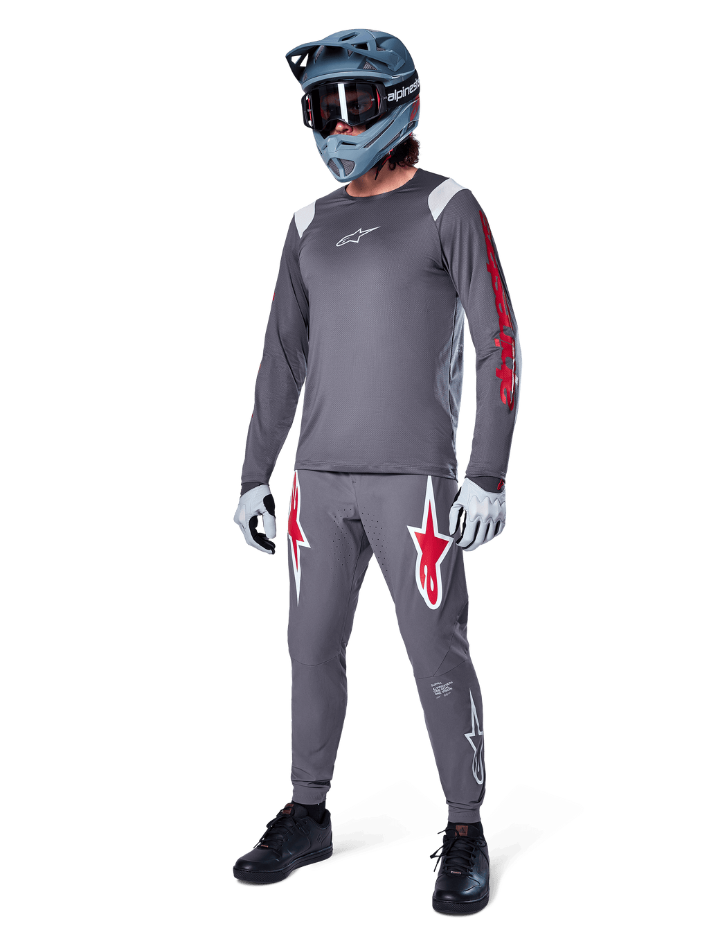 Jersey de manga larga Alpinestars A-Supra Astar en gris oscuro, con detalles de marca rojos en las mangas y logotipo blanco en el pecho, usado por un ciclista con pantalones grises a juego, casco integral gris azulado y guantes de alto rendimiento, equipamiento coordinado de MTB para gravity.
