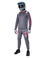 Jersey de manga larga Alpinestars A-Supra Astar en gris oscuro, con detalles de marca rojos en las mangas y logotipo blanco en el pecho, usado por un ciclista con pantalones grises a juego, casco integral gris azulado y guantes de alto rendimiento, equipamiento coordinado de MTB para gravity.