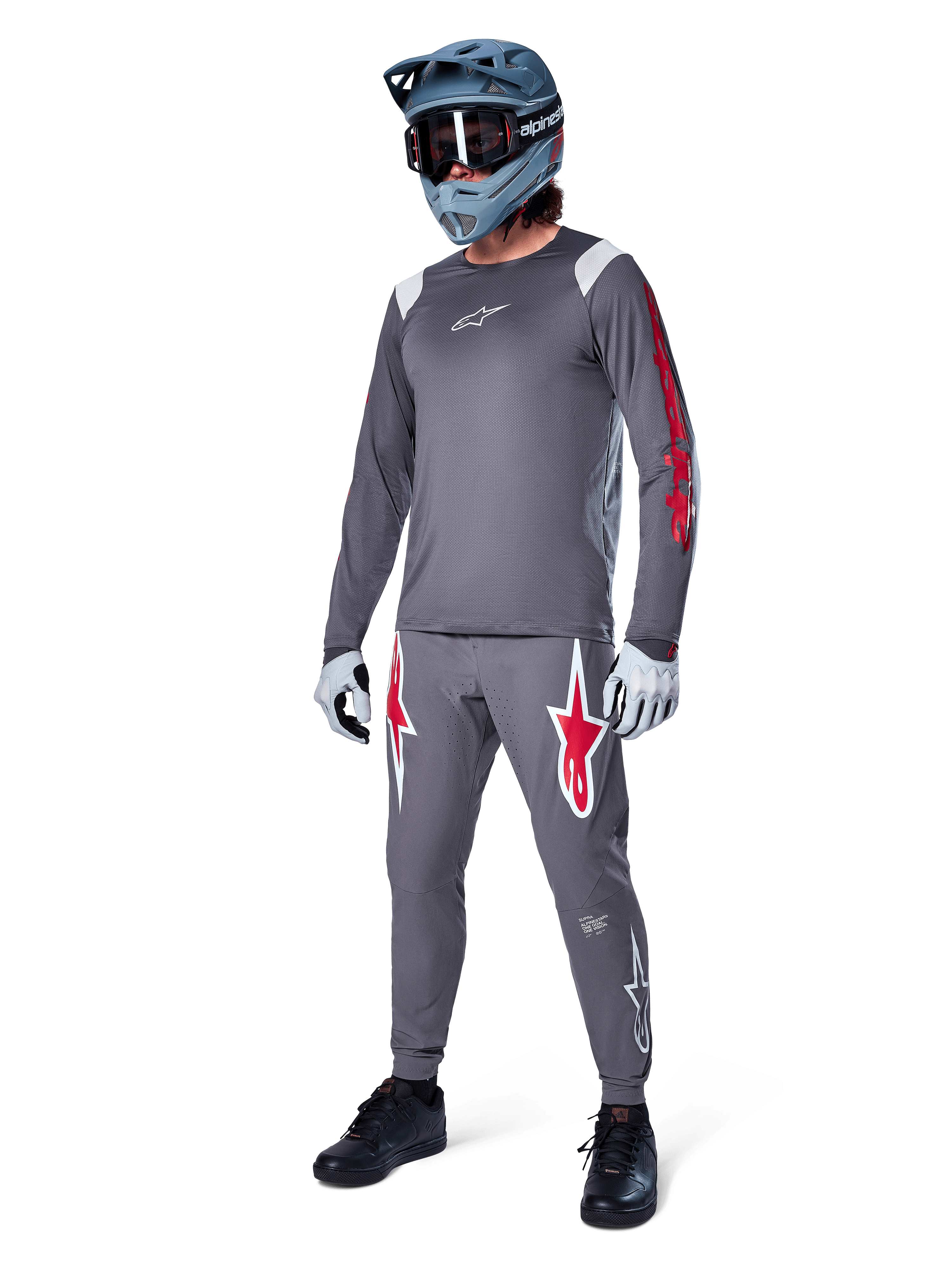 Jersey de manga larga Alpinestars A-Supra Astar en gris oscuro, con detalles de marca rojos en las mangas y logotipo blanco en el pecho, usado por un ciclista con pantalones grises a juego, casco integral gris azulado y guantes de alto rendimiento, equipamiento coordinado de MTB para gravity.