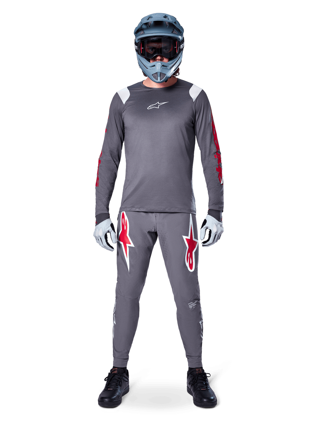 Ciclista de MTB con jersey de manga larga Alpinestars A-Supra Astar, gris oscuro con detalles rojos y logotipo blanco en el pecho, pantalones grises a juego con grandes gráficos de estrellas rojas, casco integral azul con gafas, guantes gris claro y zapatillas negras, conjunto de equipamiento profesional para bicicleta de montaña