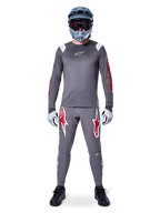 Ciclista de MTB con jersey de manga larga Alpinestars A-Supra Astar, gris oscuro con detalles rojos y logotipo blanco en el pecho, pantalones grises a juego con grandes gráficos de estrellas rojas, casco integral azul con gafas, guantes gris claro y zapatillas negras, conjunto de equipamiento profesional para bicicleta de montaña