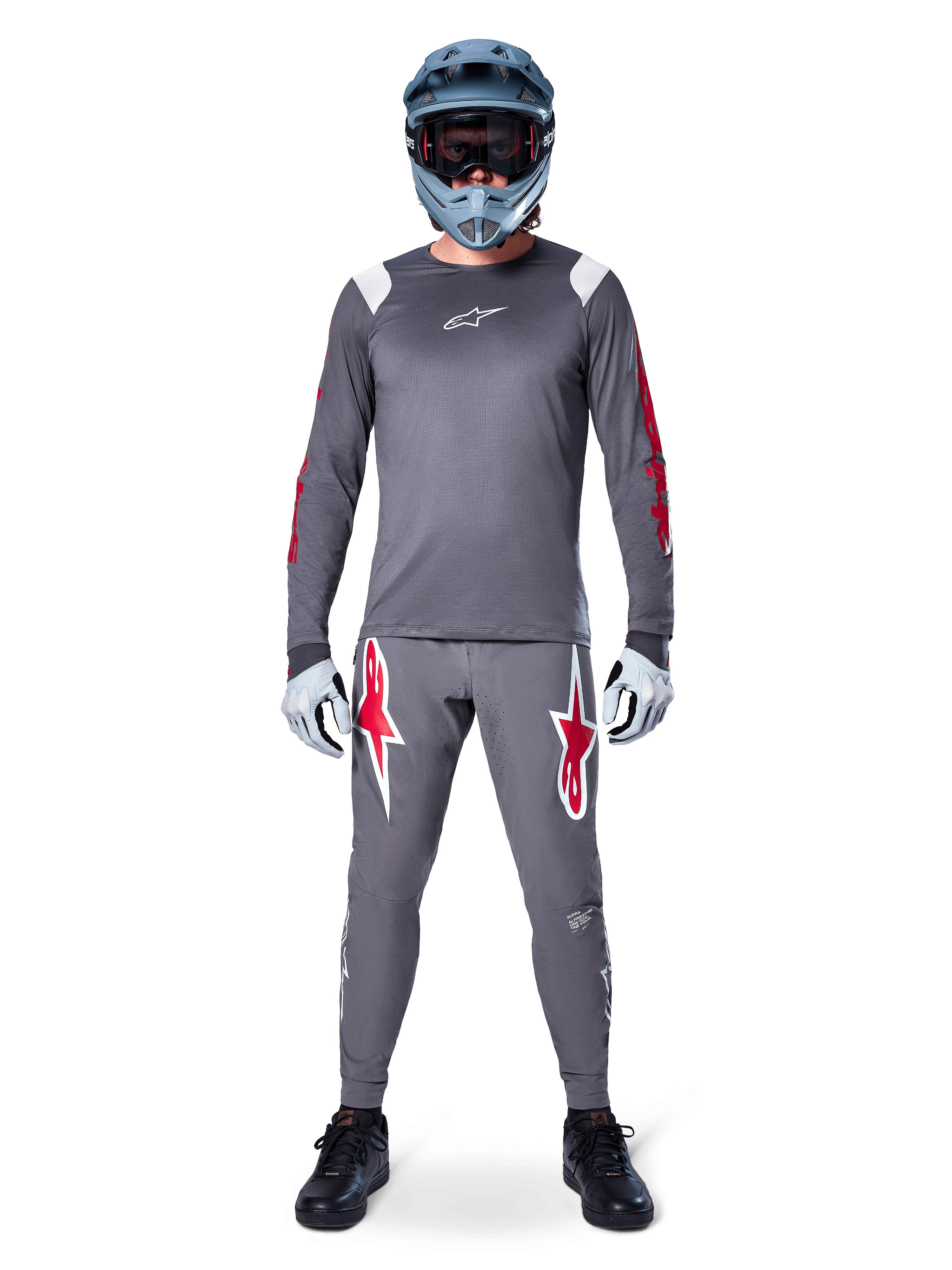 Ciclista de MTB con jersey de manga larga Alpinestars A-Supra Astar, gris oscuro con detalles rojos y logotipo blanco en el pecho, pantalones grises a juego con grandes gráficos de estrellas rojas, casco integral azul con gafas, guantes gris claro y zapatillas negras, conjunto de equipamiento profesional para bicicleta de montaña