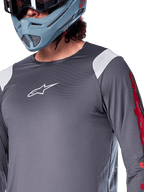 Jersey de manga larga Alpinestars A-Supra Astar, gris oscuro con paneles blancos en los hombros y gráficos rojos en las mangas, usado con un casco integral gris azulado claro, equipamiento de alto rendimiento para bicicleta de montaña