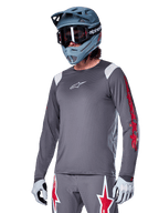 Alpinestars A-Supra Astar Jersey de manga larga, jersey para ciclismo de montaña MTB, gris oscuro con detalles rojos y blancos, ajuste de rendimiento, usado con pantalones grises a juego con gráficos de estrellas rojas y blancas, equipo completo de ciclismo todoterreno con casco integral y guantes