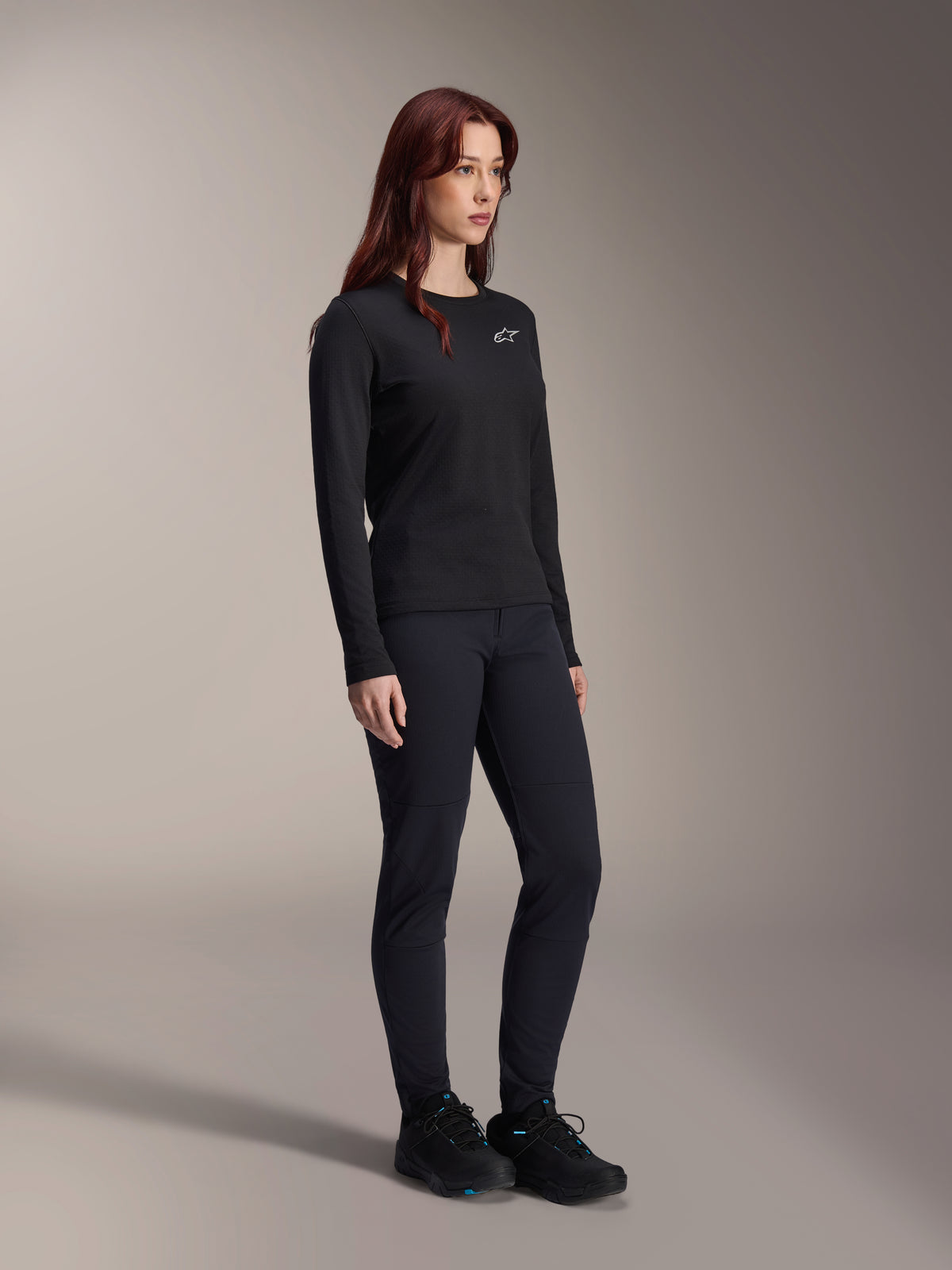 Mujer vistiendo el Jersey Térmico Alpinestars Stella A-Dura, jersey de MTB de manga larga en color negro con logotipo blanco en el pecho, combinado con pantalones negros a juego y calzado deportivo, retrato de estudio de ropa de ciclismo de alto rendimiento