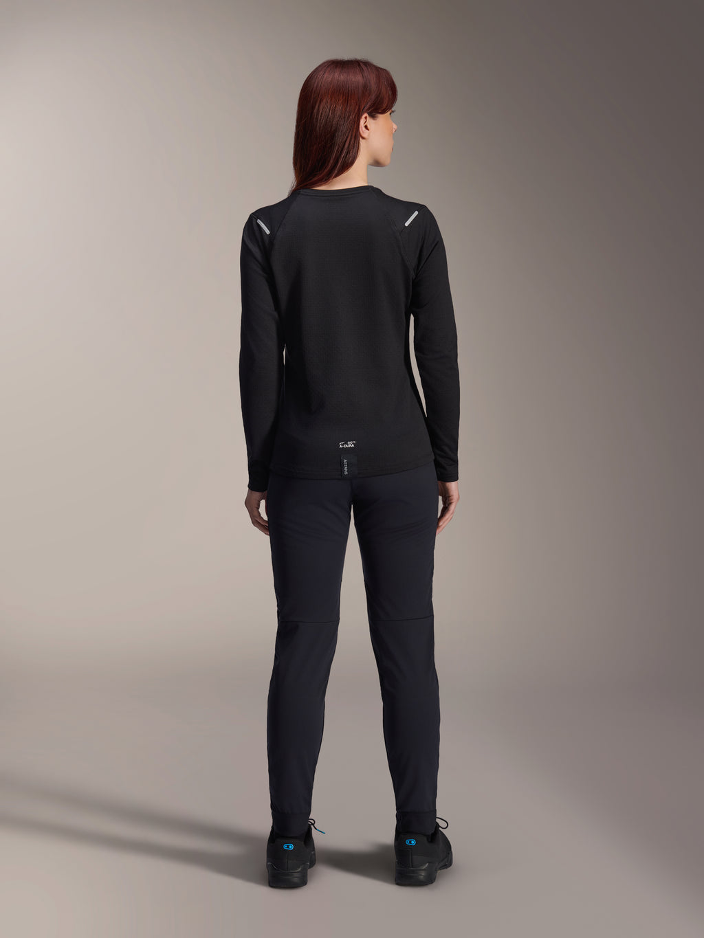 Jersey térmico Alpinestars Stella A-Dura para mujer, vista trasera, negro, jersey de ciclismo de manga larga con vellón aislante, usado con pantalones y zapatillas negras a juego, con pequeño logotipo A-Dura en la parte inferior de la espalda y detalles reflectantes en los hombros, diseñado para ciclismo de montaña femenino
