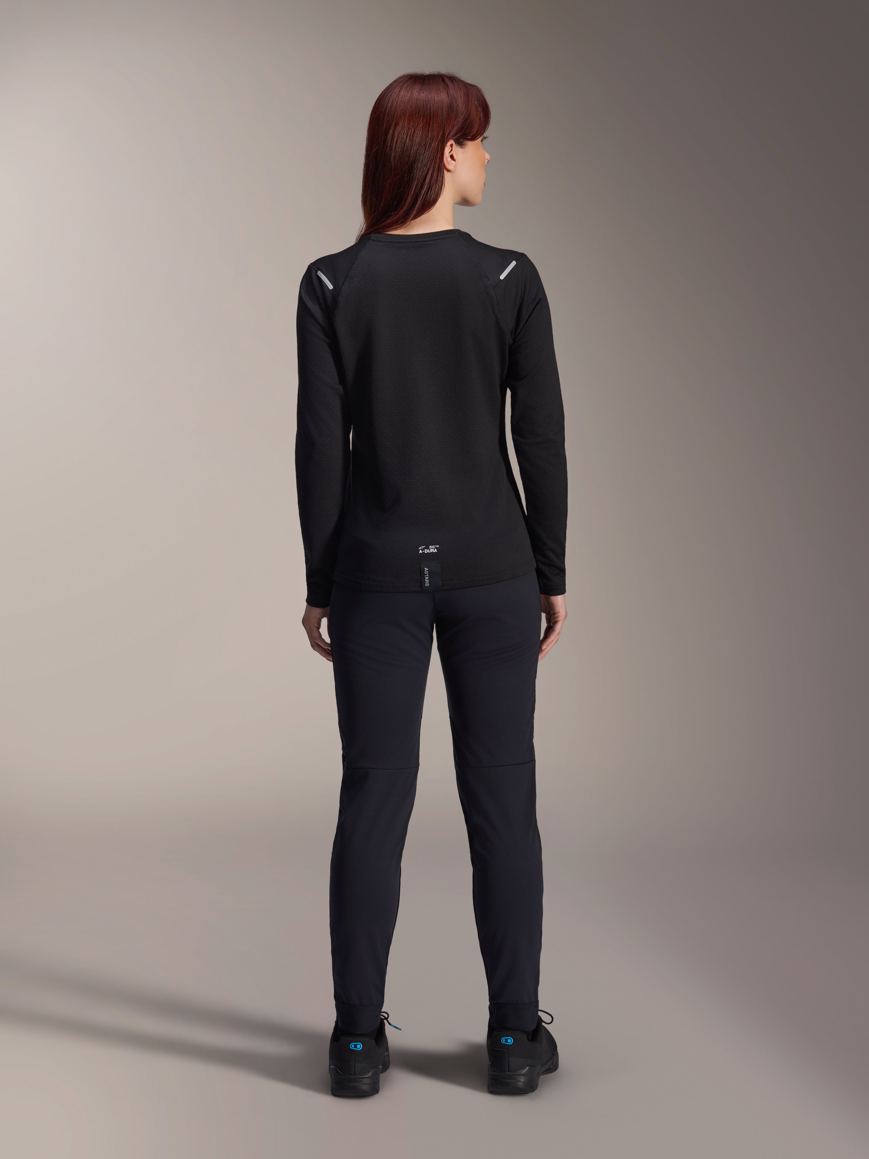 Jersey térmico Alpinestars Stella A-Dura para mujer, vista trasera, negro, jersey de ciclismo de manga larga con vellón aislante, usado con pantalones y zapatillas negras a juego, con pequeño logotipo A-Dura en la parte inferior de la espalda y detalles reflectantes en los hombros, diseñado para ciclismo de montaña femenino