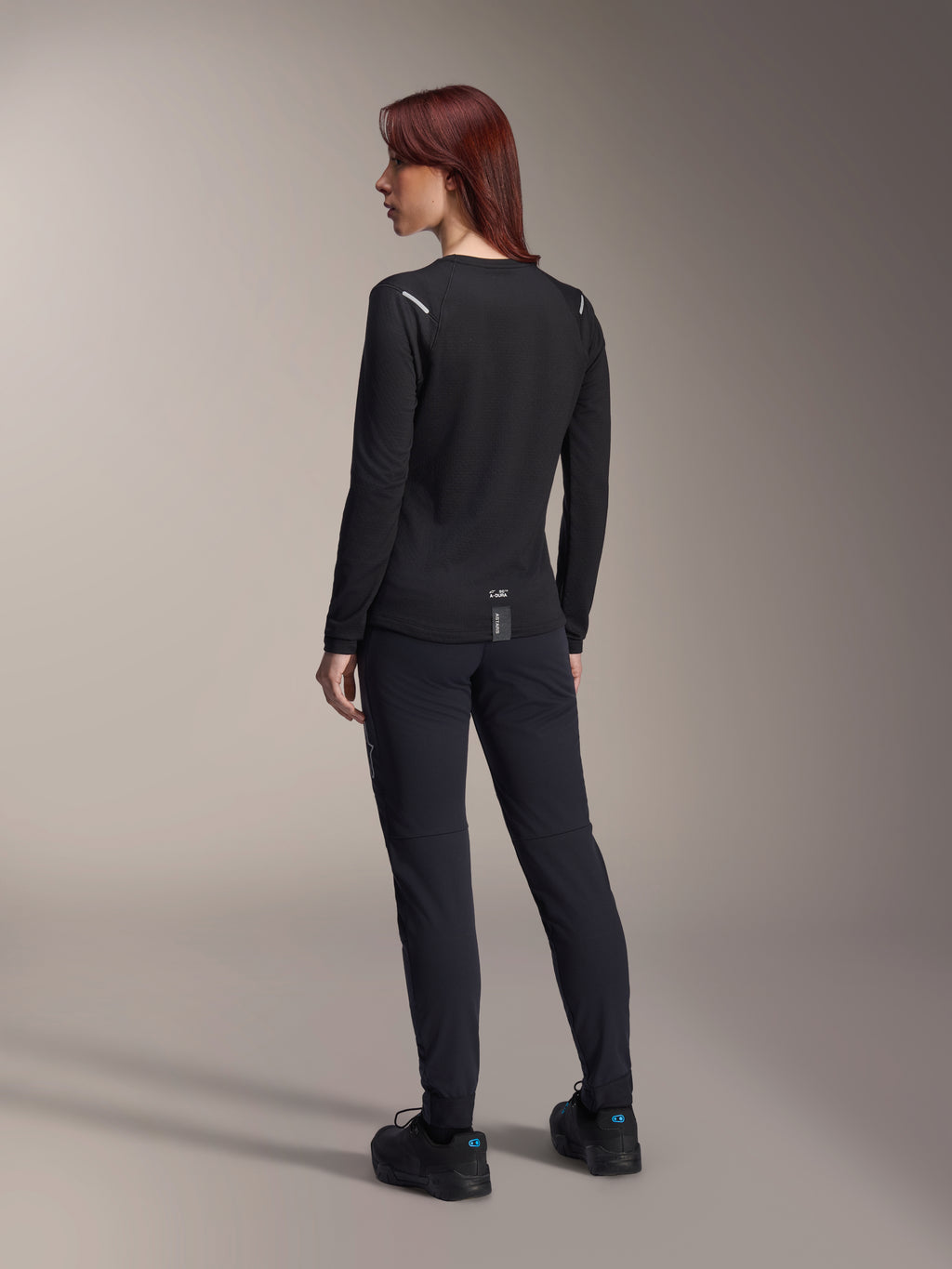 Alpinestars Stella A-Dura Thermal Jersey para mujer, jersey de MTB de manga larga en color negro, vista trasera que muestra detalles reflectantes en los hombros y parte trasera extendida, usado con pantalones y calzado negros, equipamiento de alto rendimiento para ciclismo de montaña.