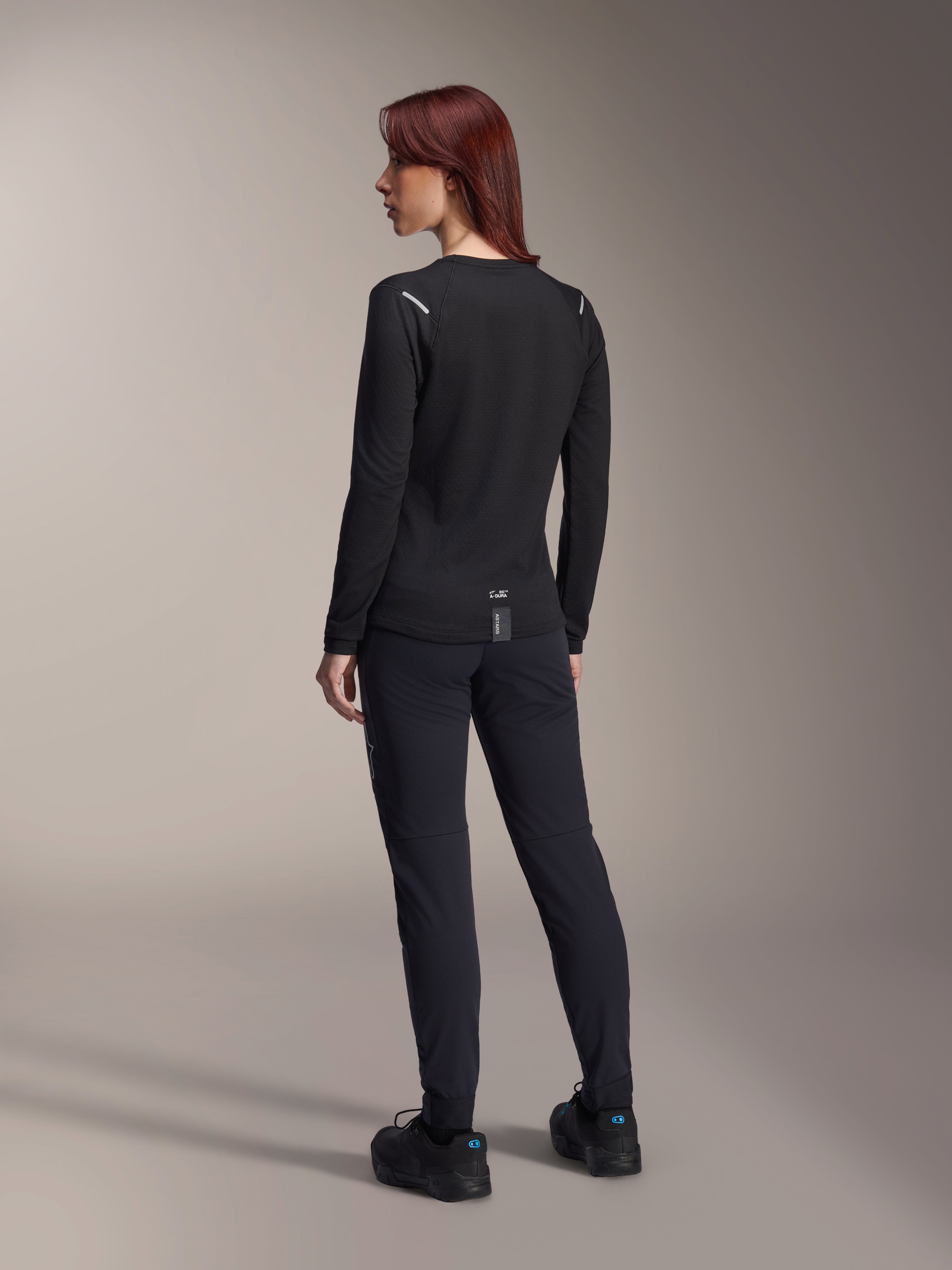 Alpinestars Stella A-Dura Thermal Jersey para mujer, jersey de MTB de manga larga en color negro, vista trasera que muestra detalles reflectantes en los hombros y parte trasera extendida, usado con pantalones y calzado negros, equipamiento de alto rendimiento para ciclismo de montaña.