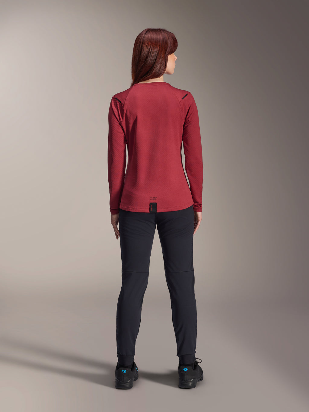 Mujer con el Jersey Térmico Stella A-Dura de Alpinestars, color borgoña, vista trasera, jersey de ciclismo de manga larga con aislamiento y cola trasera extendida, combinado con pantalones negros y calzado de ciclismo negro, ropa para ciclismo de montaña.