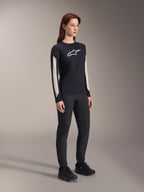 Woman wearing Alpinestars MTB gear, black Stella A-Dura Elite Jersey with white chest logo and sleeve panels, black Stella A-Dura Pants, black cycling shoes with blue accents, full performance outfit for mountain biking