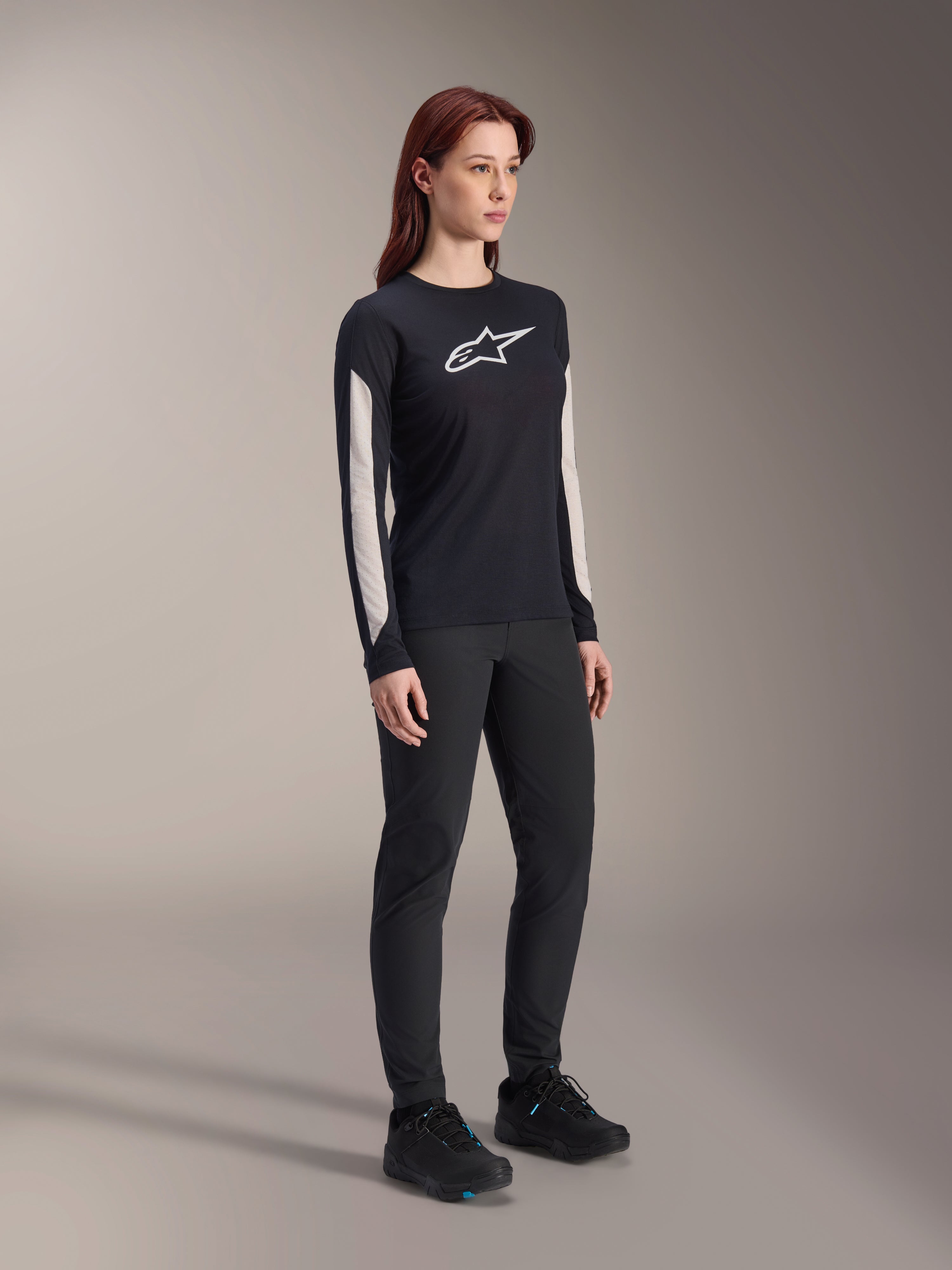 Woman wearing Alpinestars MTB gear, black Stella A-Dura Elite Jersey with white chest logo and sleeve panels, black Stella A-Dura Pants, black cycling shoes with blue accents, full performance outfit for mountain biking