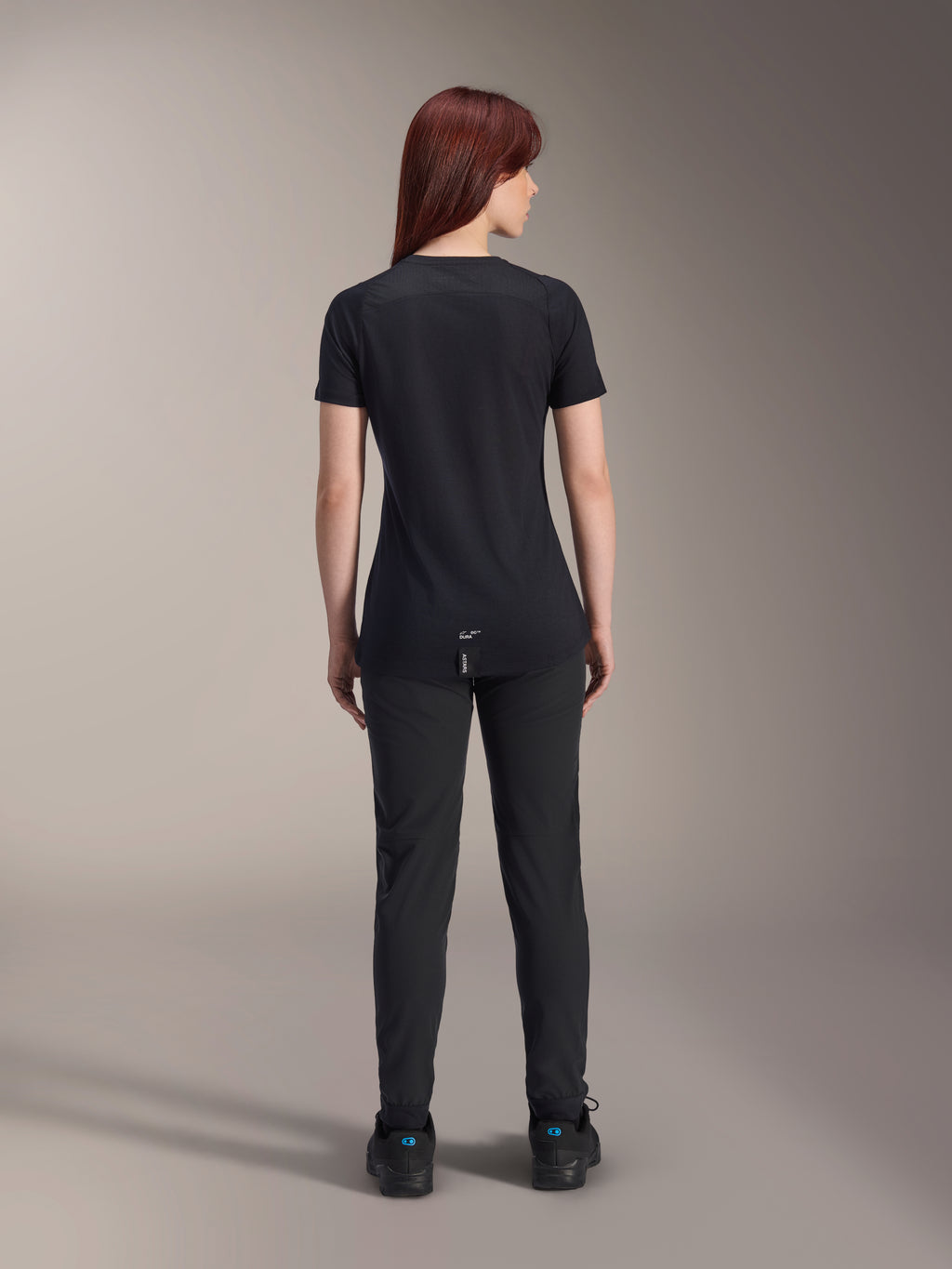 Alpinestars Jersey Stella A-Dura Dri Switch de manga corta para mujer, negro, vista trasera en una modelo, con cola más larga y paneles de malla técnica, combinado con pantalones negros, equipamiento de alto rendimiento para ciclismo MTB