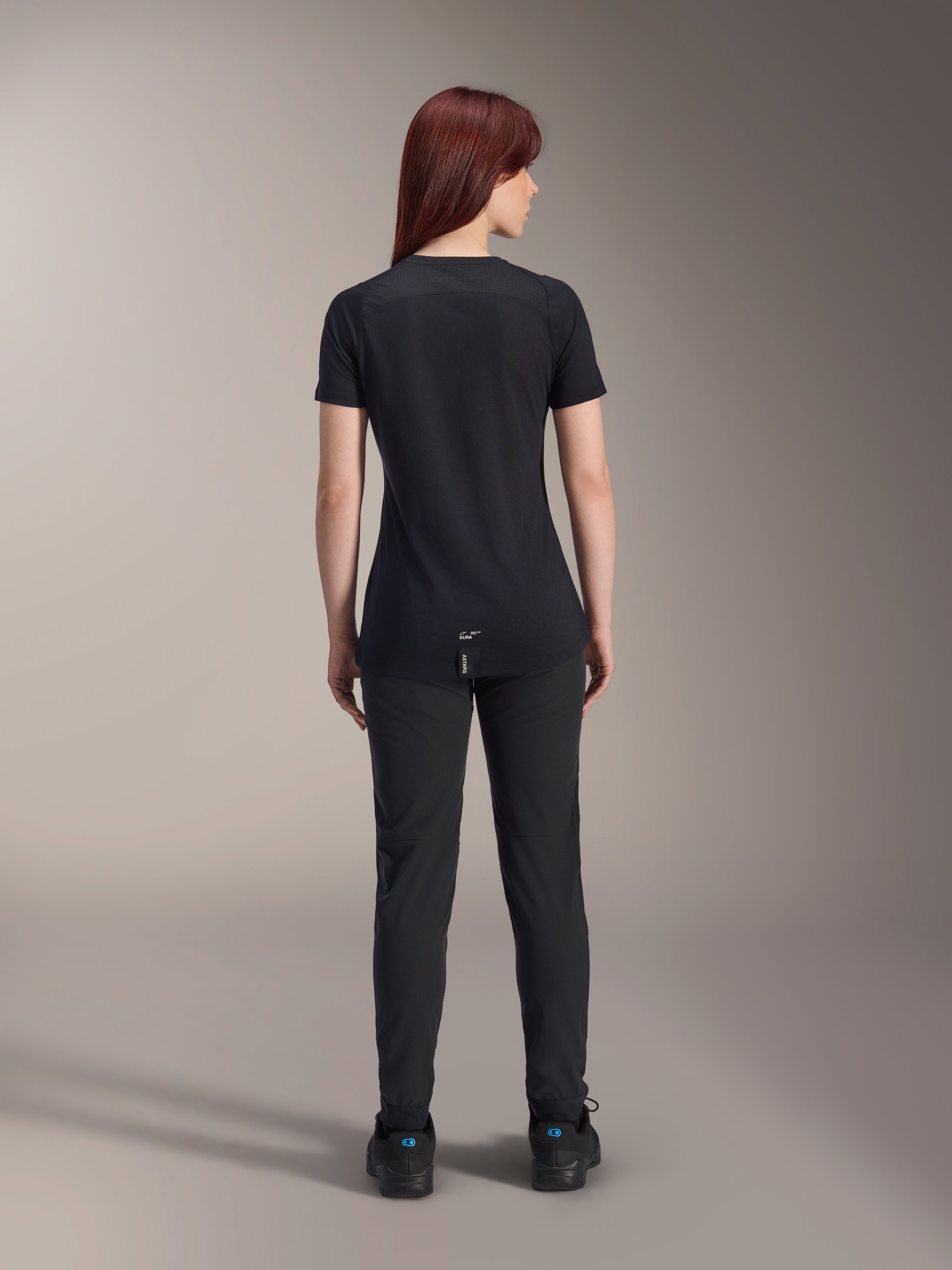 Alpinestars Jersey Stella A-Dura Dri Switch de manga corta para mujer, negro, vista trasera en una modelo, con cola más larga y paneles de malla técnica, combinado con pantalones negros, equipamiento de alto rendimiento para ciclismo MTB