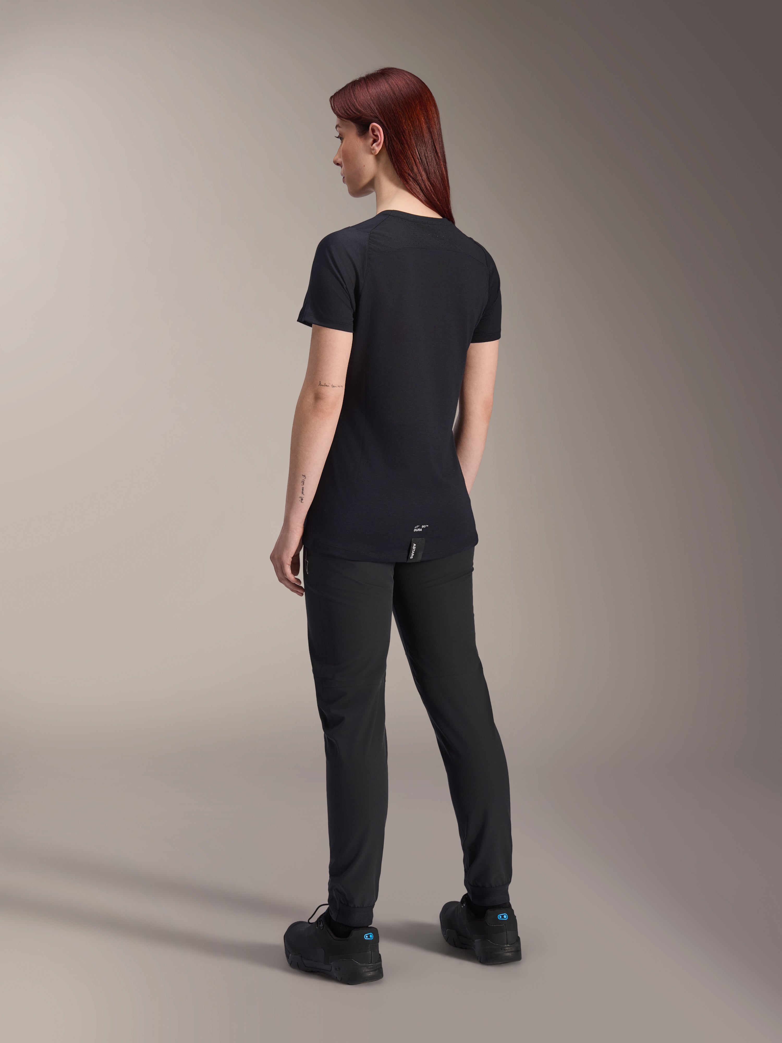 Modelo pelirroja con el jersey Alpinestars Stella A-Dura Dri Switch de manga corta en negro, vista trasera, combinado con pantalones y zapatillas de ciclismo de montaña negros, mostrando el ajuste técnico y la cola más larga para mayor cobertura.