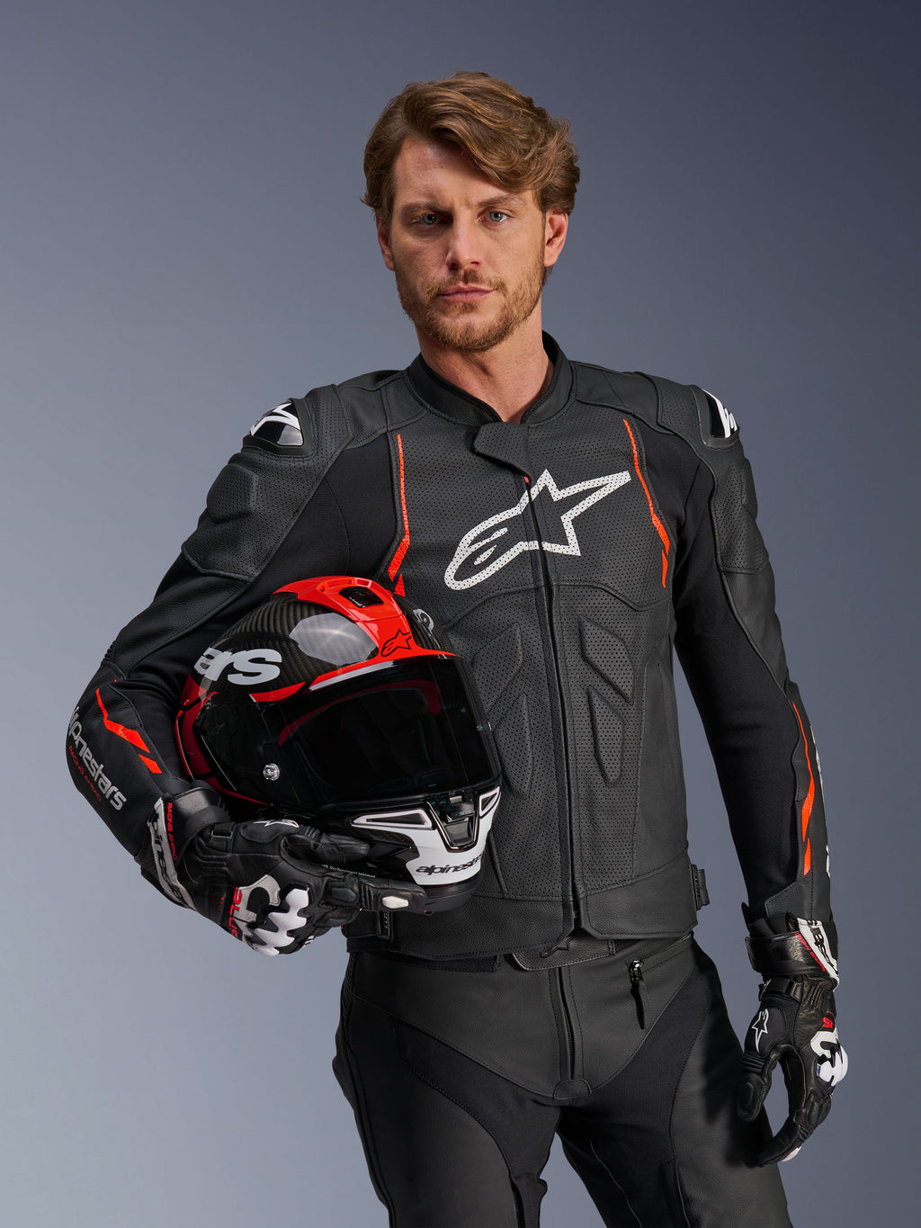 Chaqueta de cuero Alpinestars Dusk Airflow, negra con detalles en rojo flúor, usada por un modelo masculino que sostiene un casco de moto rojo y negro, con cuero perforado para una máxima ventilación y logotipo de Alpinestars en blanco, equipamiento de conducción deportiva para motocicleta