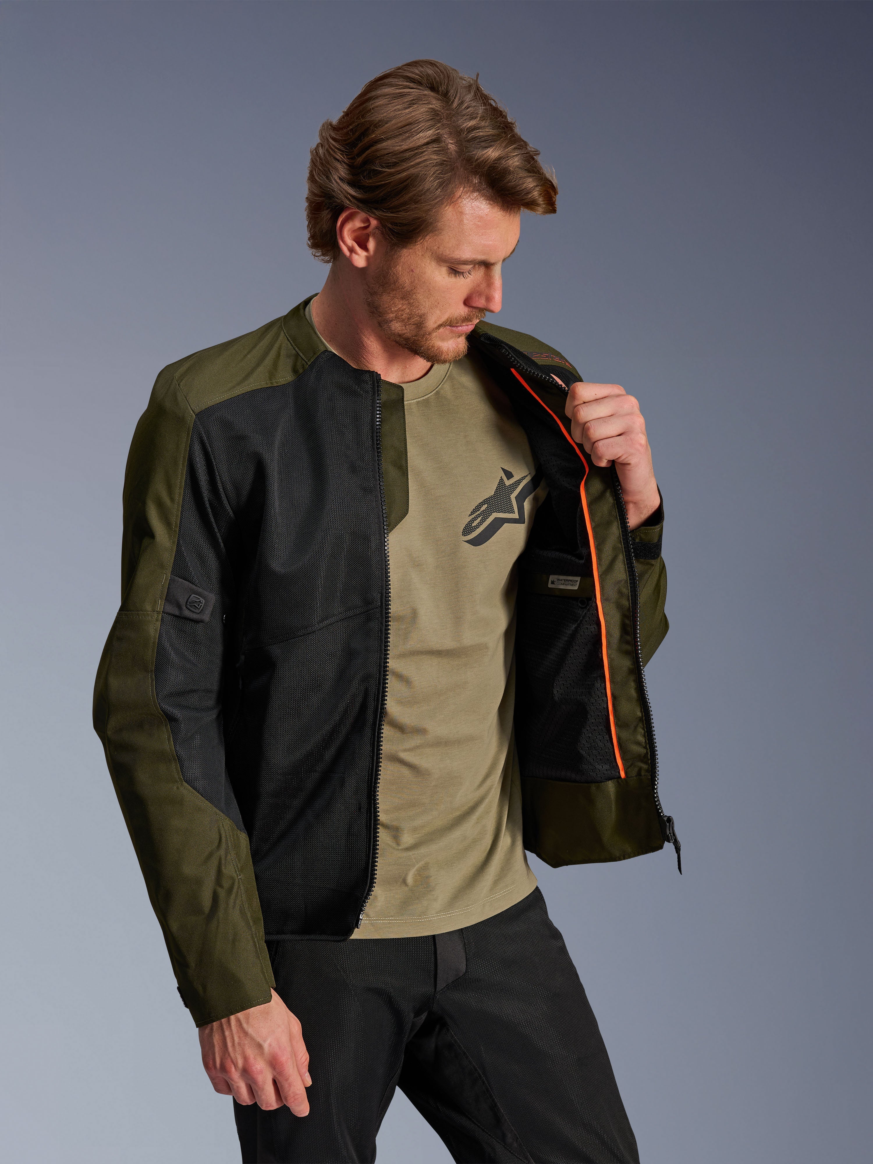 Chaqueta Alpinestars C-1 Air en caqui y negro, usada por un modelo masculino para mostrar el forro interior naranja y el compartimento impermeable, con grandes paneles de malla técnica para el flujo de aire, de estilo urbano contemporáneo combinada con una camiseta oliva a juego y pantalones negros