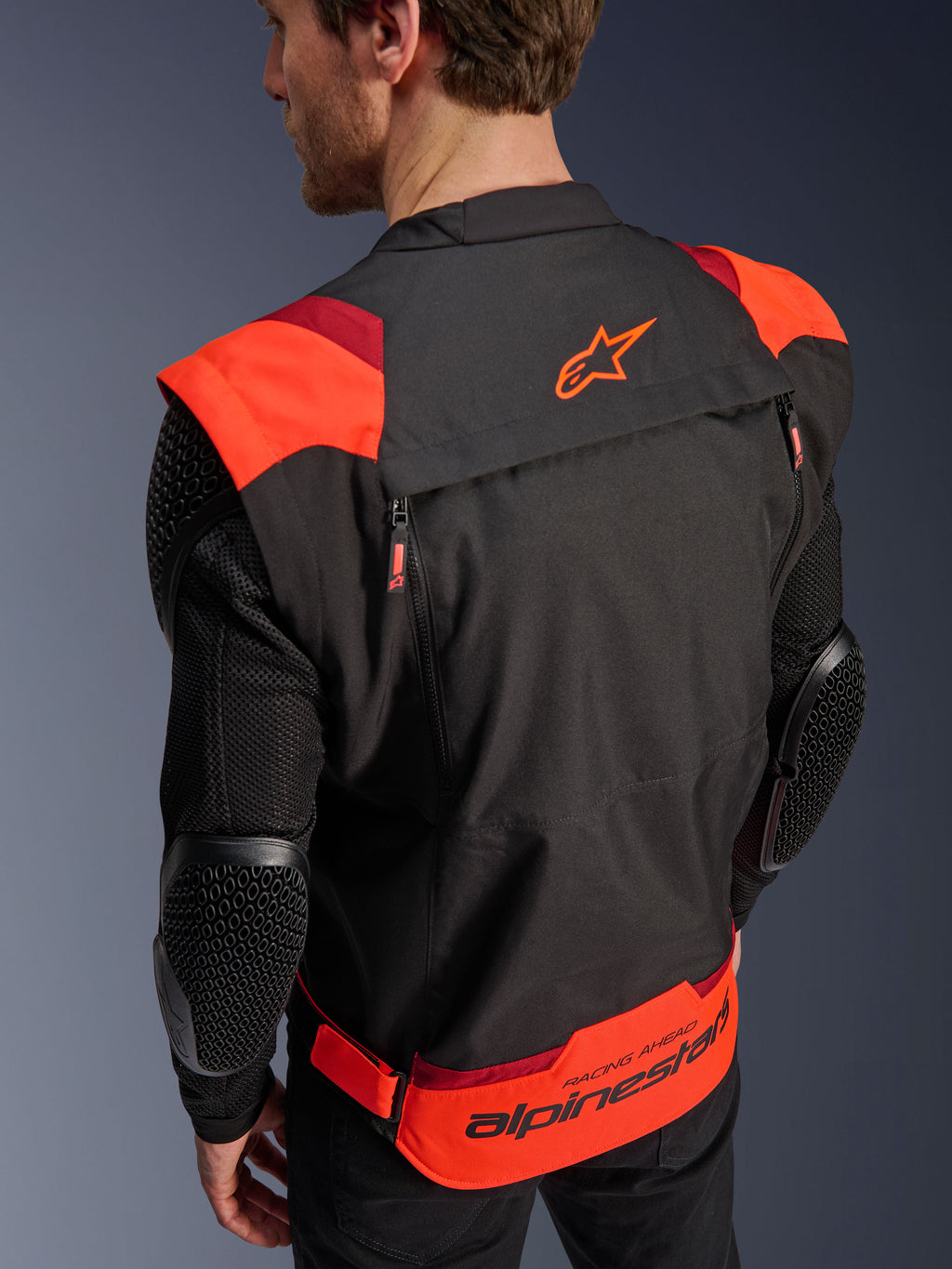 Chaqueta Alpinestars T-Stunt Air, negro, rojo flúor y rojo oscuro, vista posterior en un modelo, que muestra mangas de malla con protectores de codo con textura de panal, el logotipo de la estrella de Alpinestars en la parte superior de la espalda y una marca llamativa en la parte inferior con el texto 'RACING AHEAD', diseño modular de corte deportivo para motociclismo.