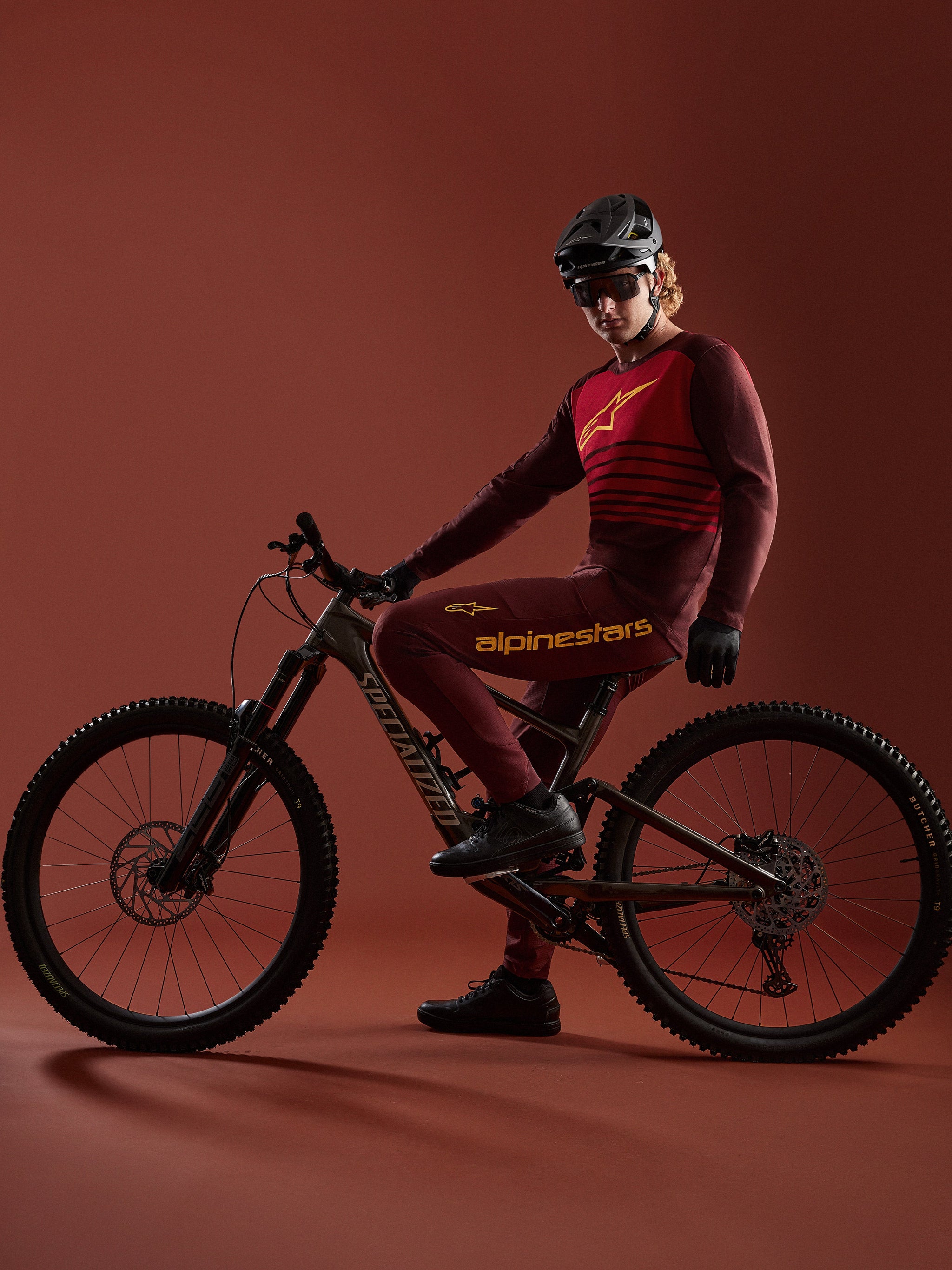 Conjunto bordeaux A-Dura Elite 2.0 de Alpinestars, ciclista de montaña con bicicleta Specialized en estudio.