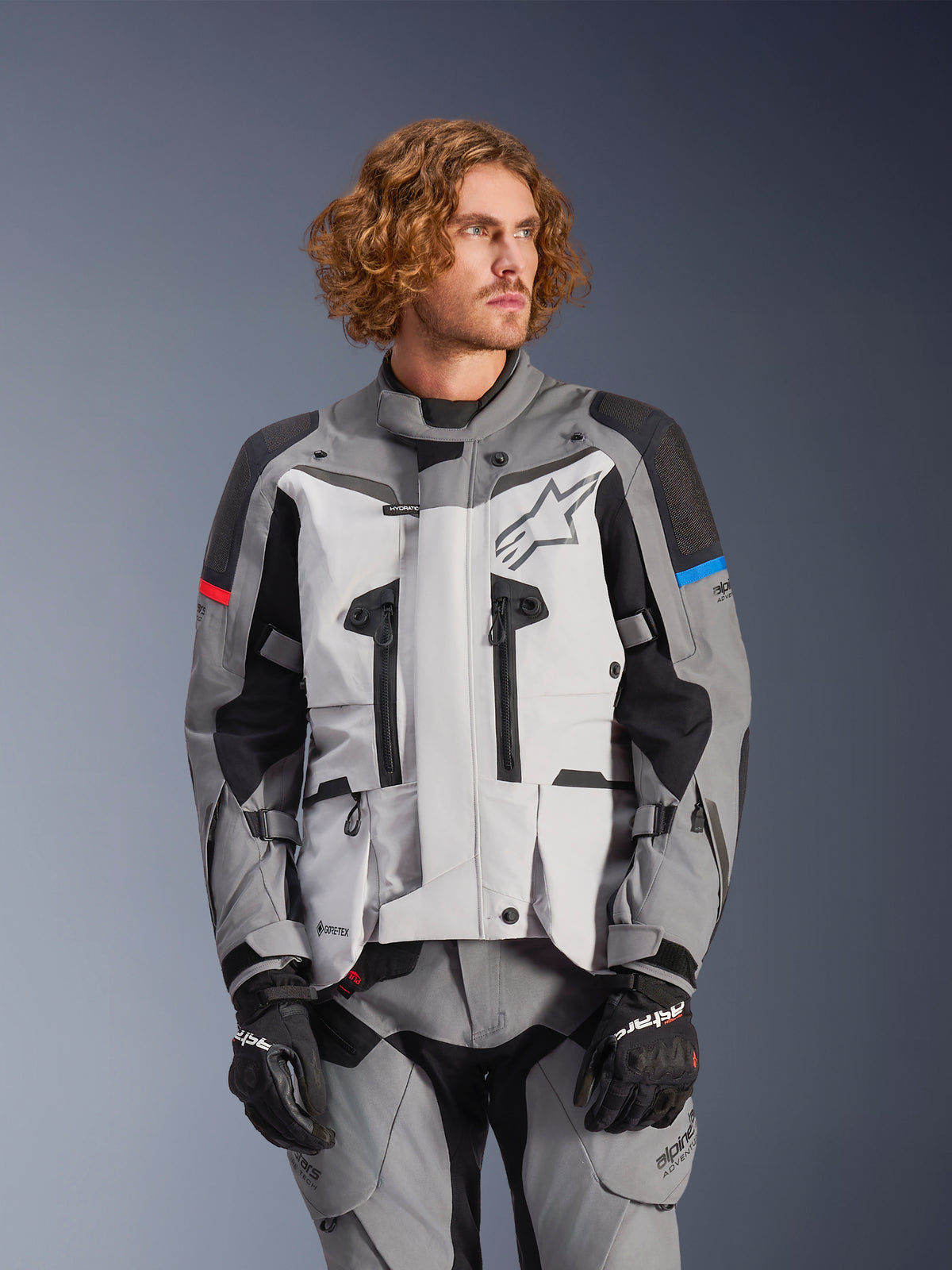 Alpinestars Boulder GORE-TEX Jacket, chaqueta de moto para turismo, gris y negra con detalles rojos y azules en las mangas, impermeable y transpirable, con cremalleras y logo de estrella, persona con pantalones grises y guantes negros a juego