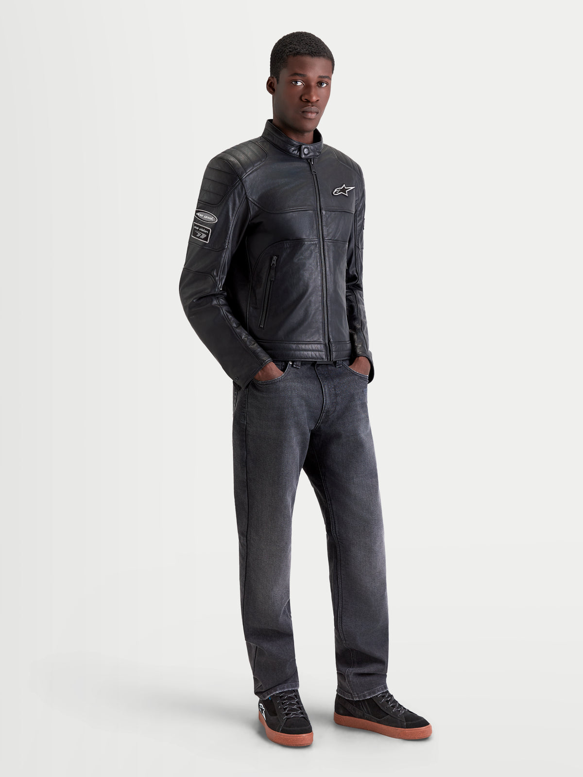 Alpinestars Revern chaqueta de cuero para moto urbana para hombre, color negro, vista frontal en modelo con vaqueros oscuros y zapatos de moto, diseño de estilo heritage con construcción de cuero vacuno de plena flor