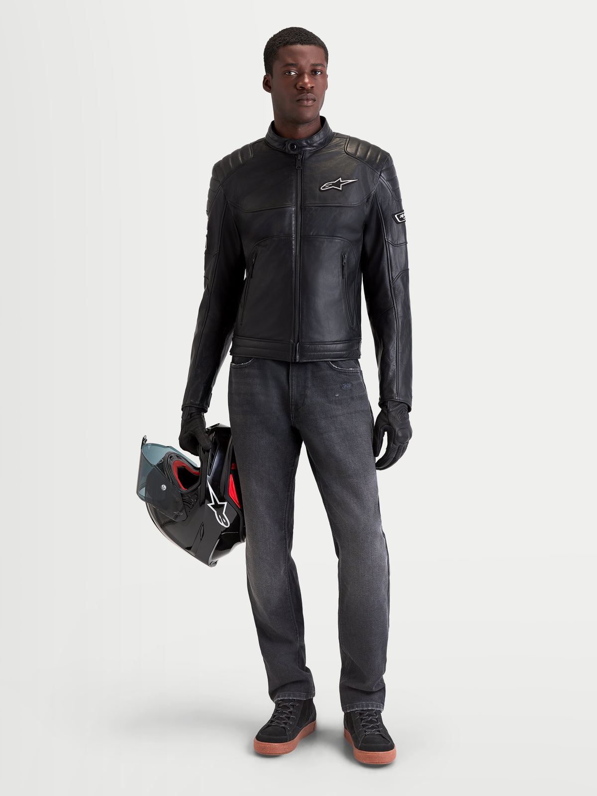 Alpinestars Revern Chaquetas de cuero, chaqueta de moto urbana para hombre, negra, vista frontal en un modelo que lleva vaqueros oscuros y sostiene un casco, con construcción de cuero bovino envejecido y estilo motero heritage