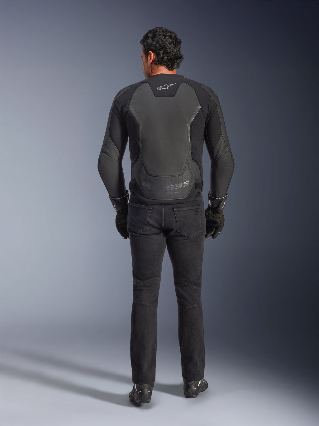 Chaqueta de cuero Alpinestars GP Force V2, negra, vista trasera, chaqueta de moto deportiva premium con construcción de cuero y textil, usada con vaqueros de moto negros y guantes, con paneles protectores en los hombros y detalles de la marca