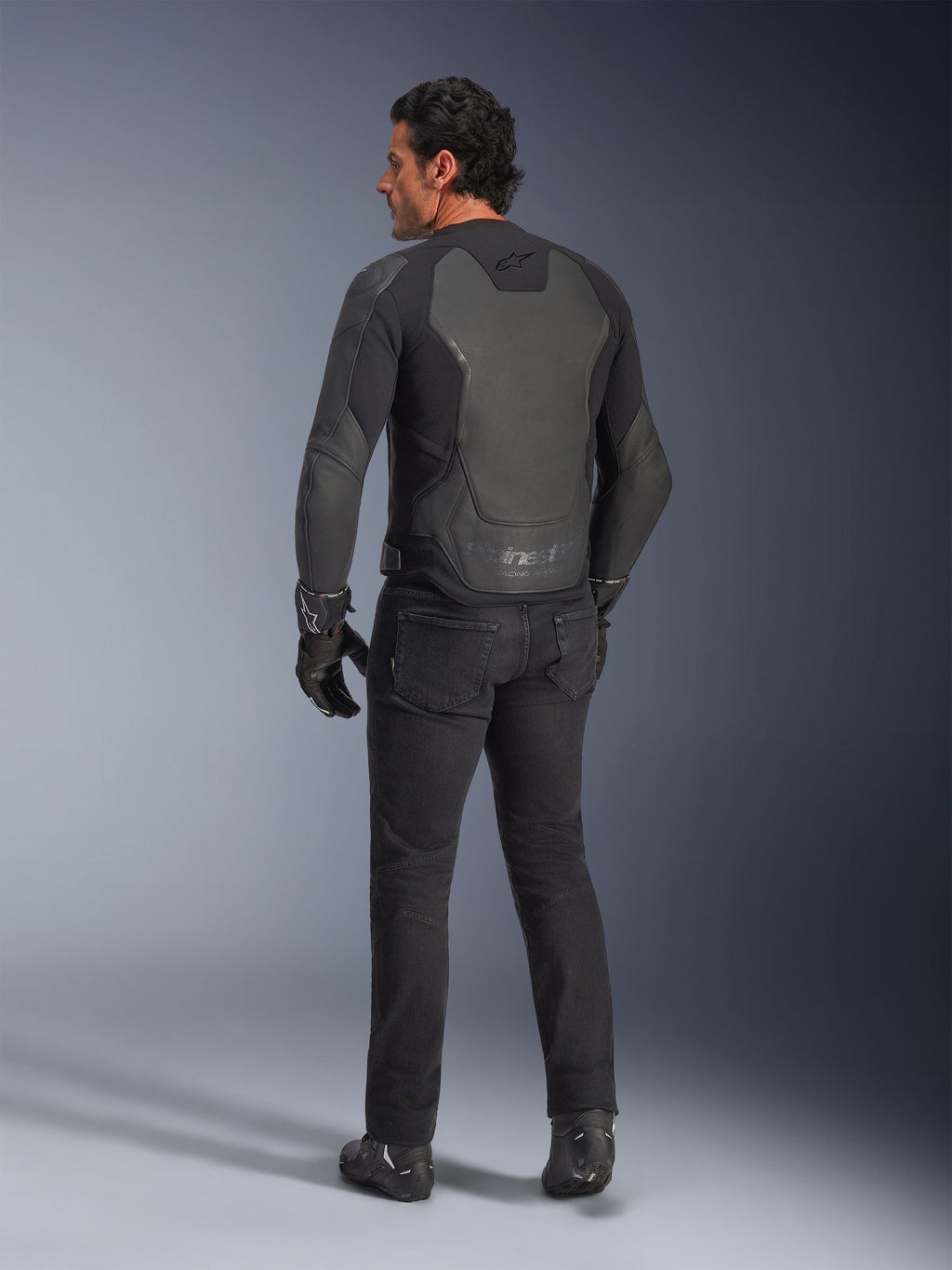 Vista trasera de un modelo vistiendo la Chaqueta de Cuero Alpinestars GP Force V2 en color Black Black, con una construcción de cuero de múltiples paneles y ajuste deportivo, combinada con pantalones de conducción, guantes y botas negros para un look completo inspirado en las carreras de motos.
