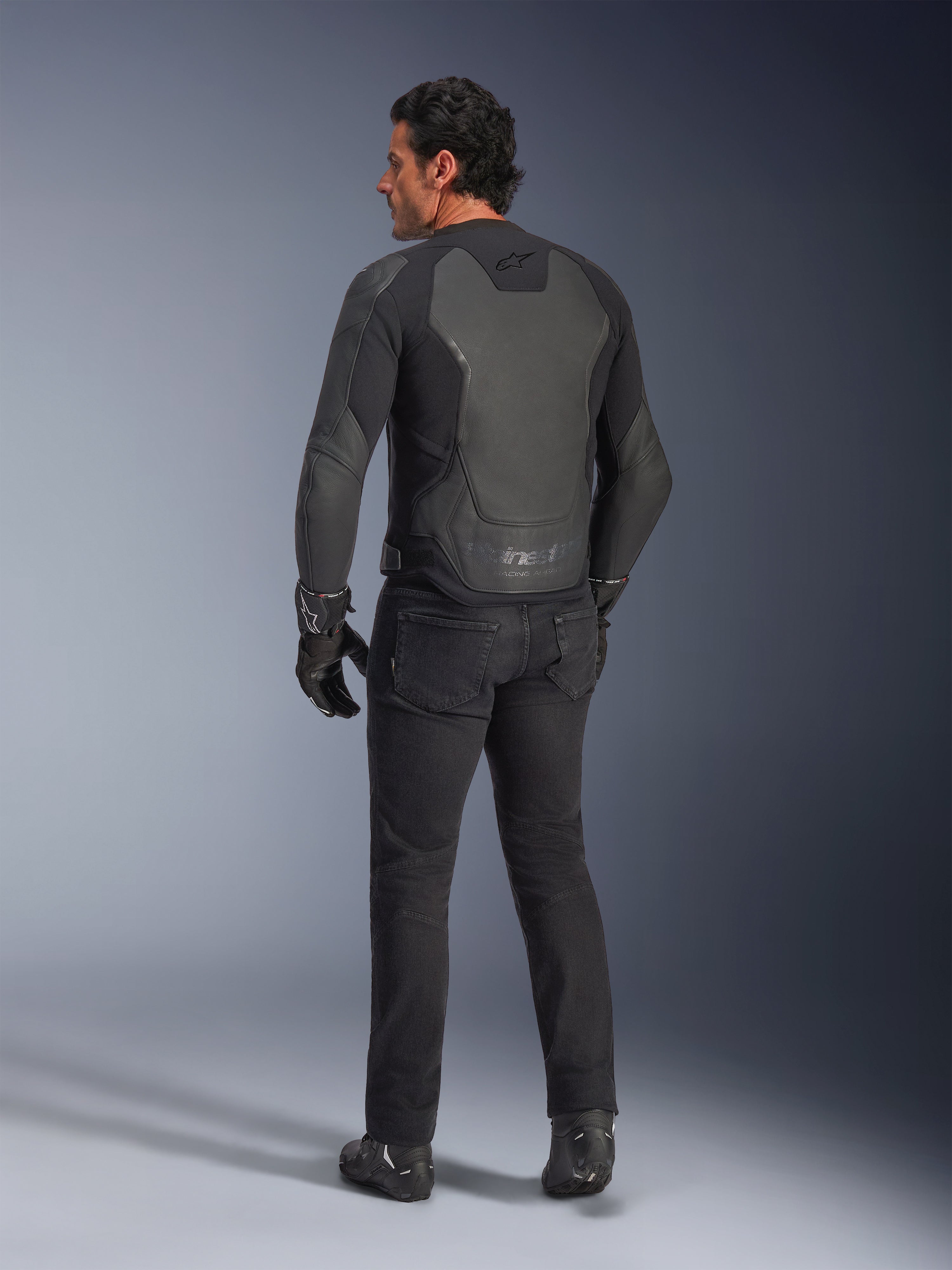 Vista trasera de un modelo vistiendo la Chaqueta de Cuero Alpinestars GP Force V2 en color Black Black, con una construcción de cuero de múltiples paneles y ajuste deportivo, combinada con pantalones de conducción, guantes y botas negros para un look completo inspirado en las carreras de motos.