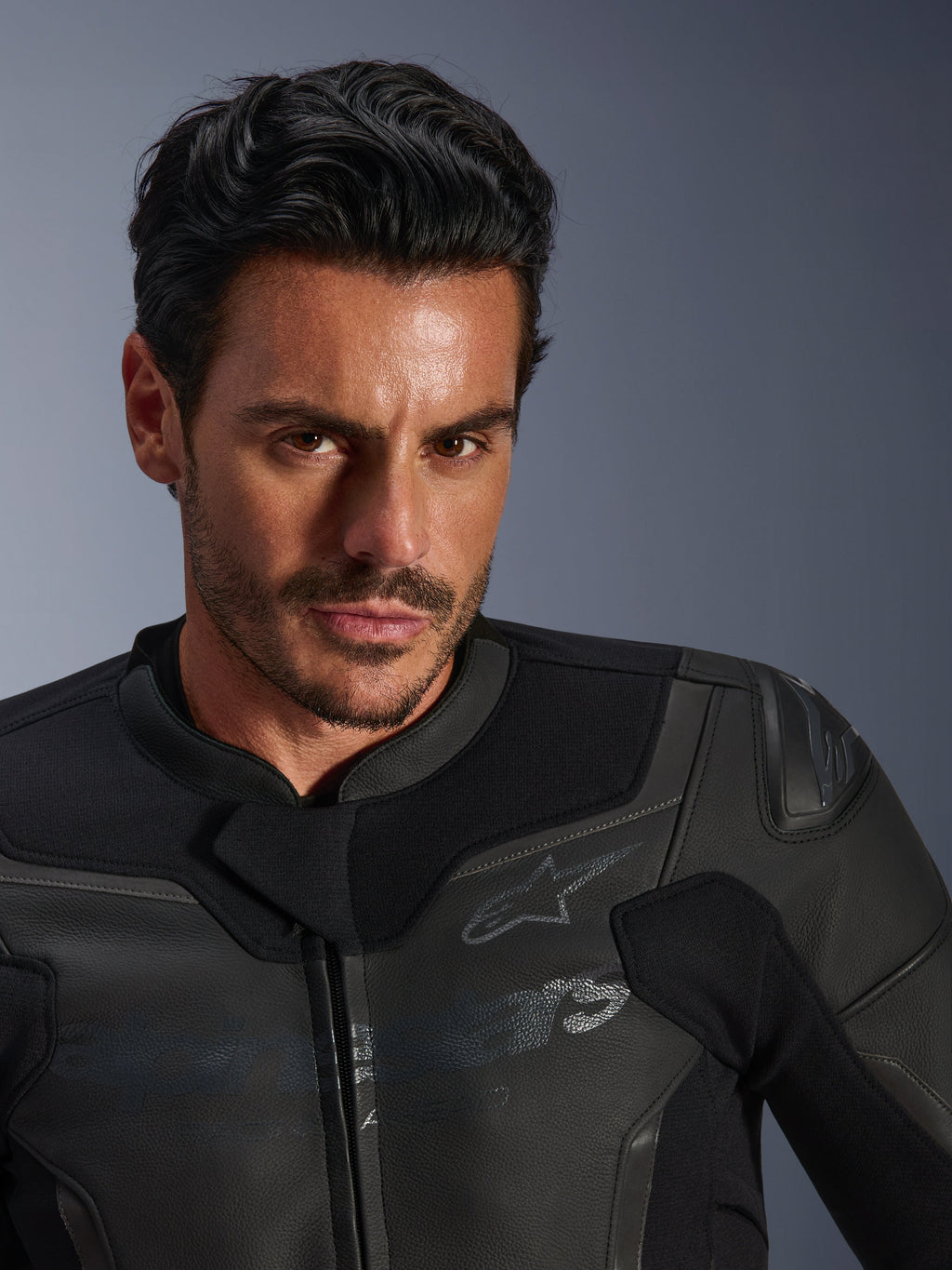 Retrato en primer plano de un modelo masculino vistiendo la chaqueta de cuero Alpinestars GP Force V2, negra, con confección en cuero bovino de primera calidad, paneles elásticos textiles y un sutil logotipo de la estrella de Alpinestars, diseño deportivo estilizado para conducción en carretera.